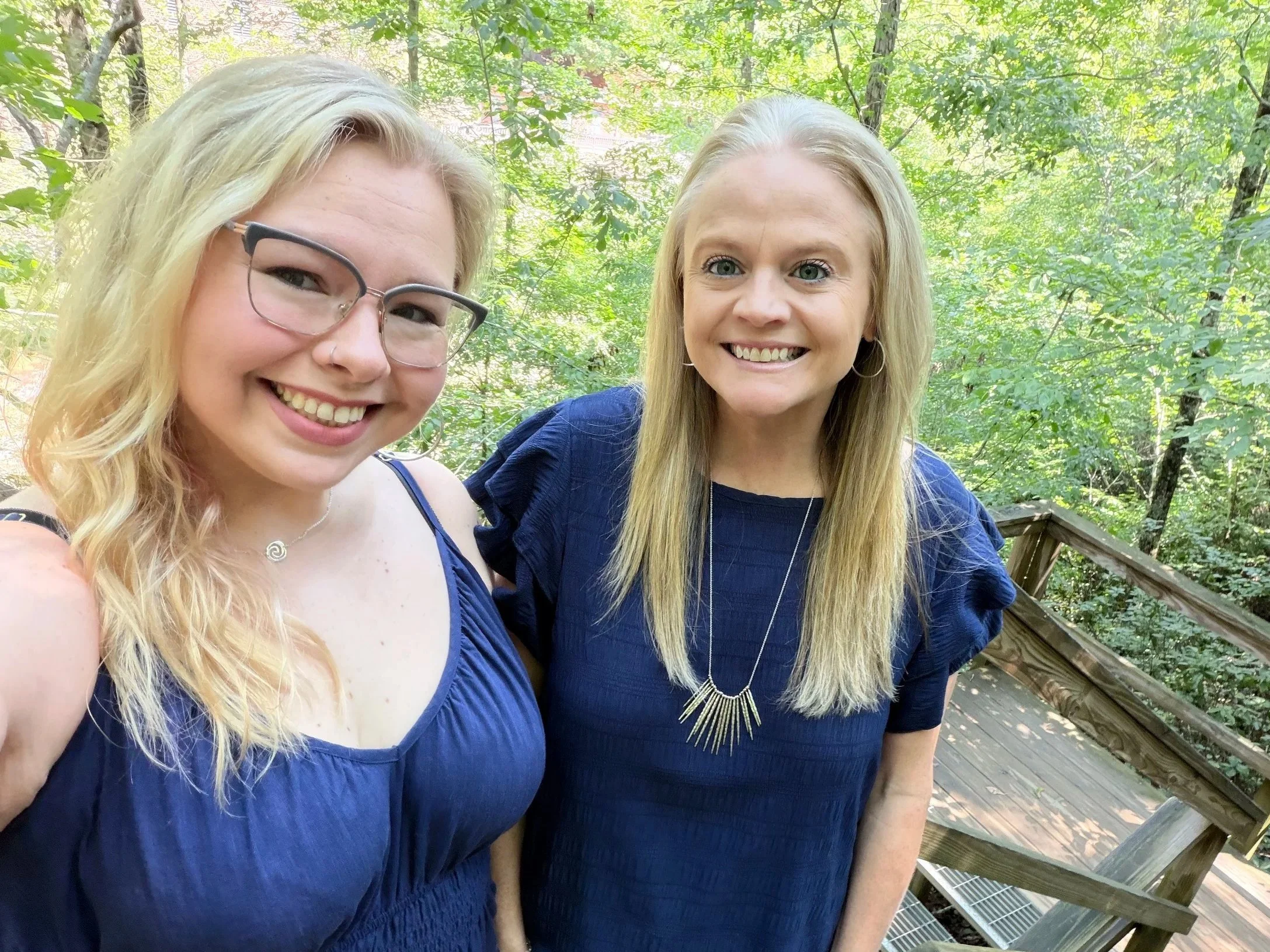 Two women smiling for a selfie outdoors in a forested area, standing on a wooden deck with green trees in the background. Both women have blonde hair and are wearing blue tops.