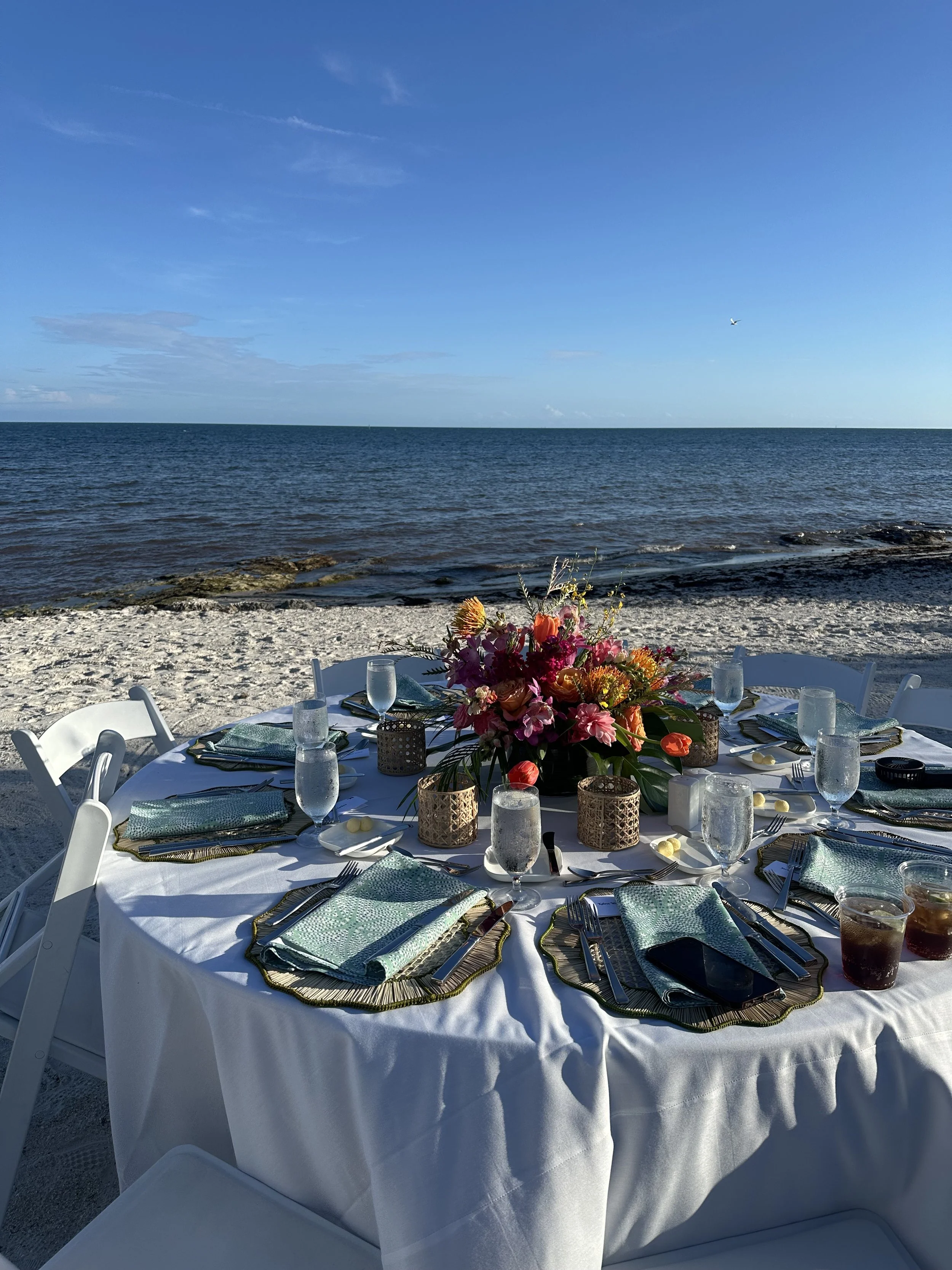 Round table set for a meal on a beach with a floral centerpiece, surrounded by white chairs, overlooking the ocean under a blue sky with a few clouds and a plane in the distance.