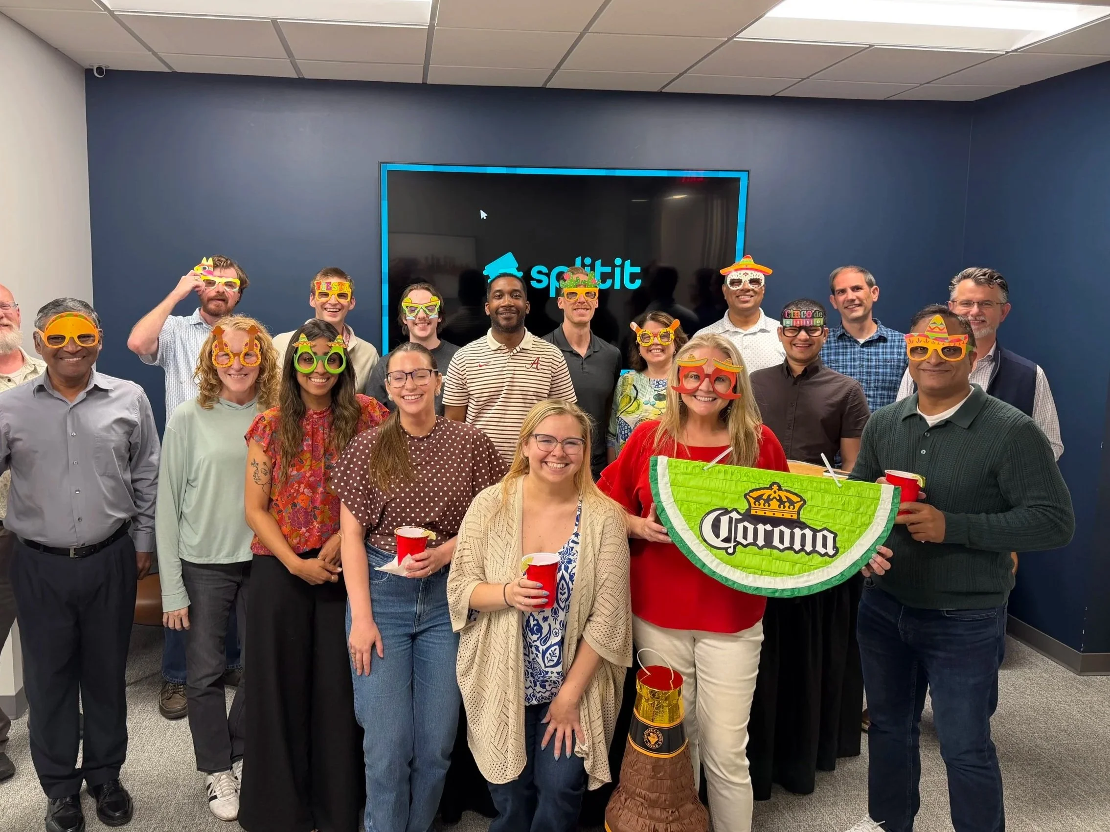 A group of people celebrating at a party, wearing colorful masks and holding red cups, with a large Corona beer pinata in the front. They are smiling and standing in an office with a blue wall and a TV screen in the background.