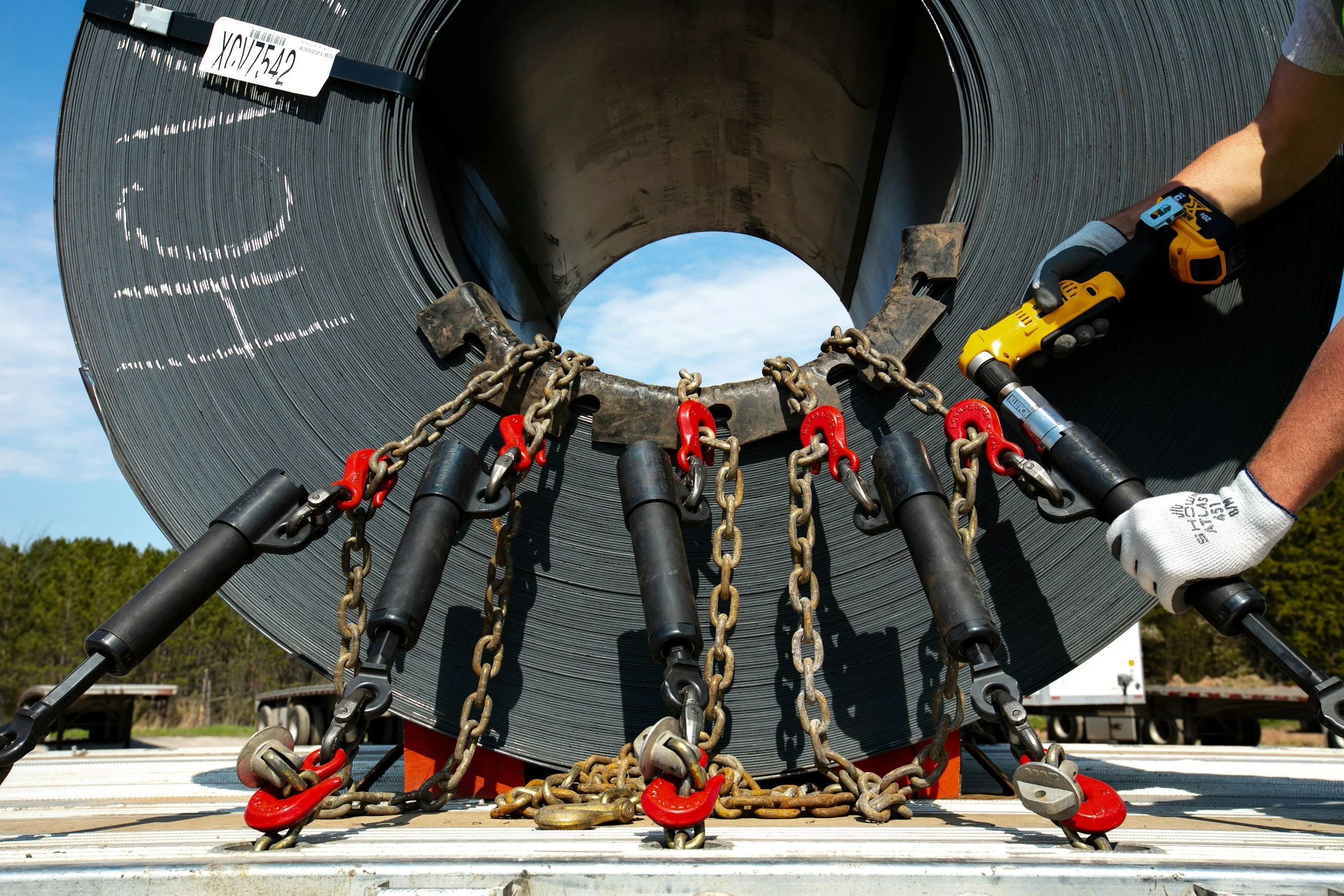 Close-up of a large black metal coil secured with chains and red hooks, with a person holding a yellow power tool working on it, outdoors on a sunny day with trees and trucks in the background.
