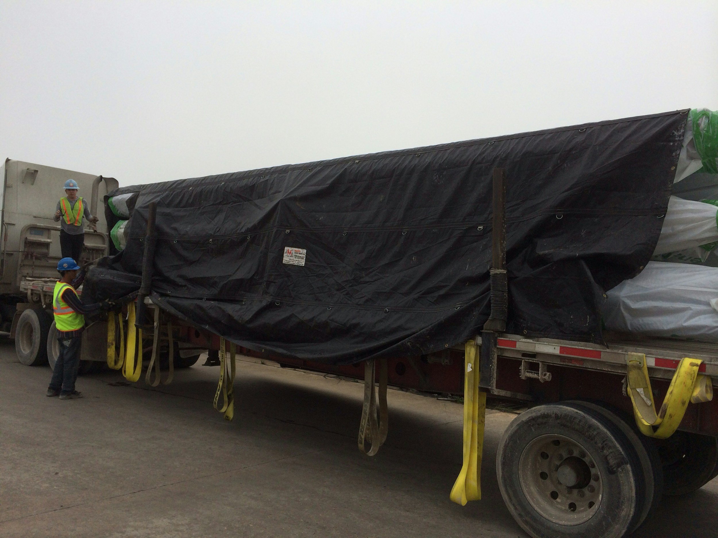 Construction workers wearing safety vests and helmets loading or unloading large wrapped equipment onto a flatbed truck during daytime.