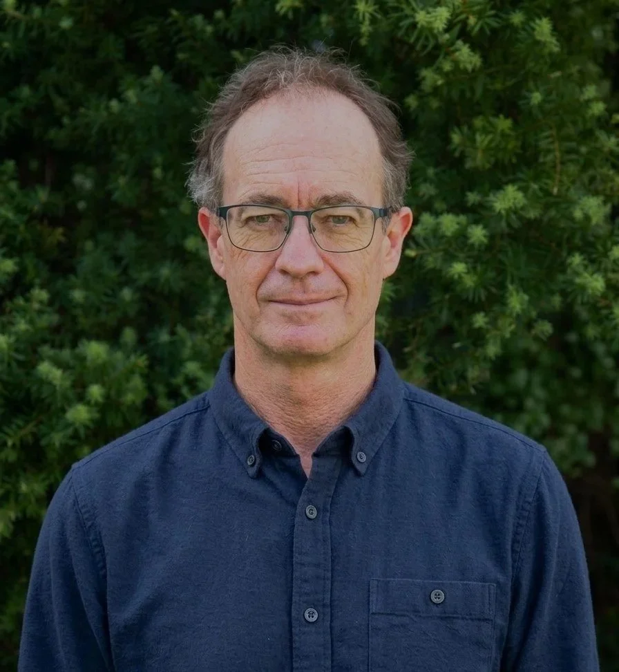 A middle-aged man with glasses and short brown hair standing outdoors in front of green foliage, wearing a navy blue button-up shirt.