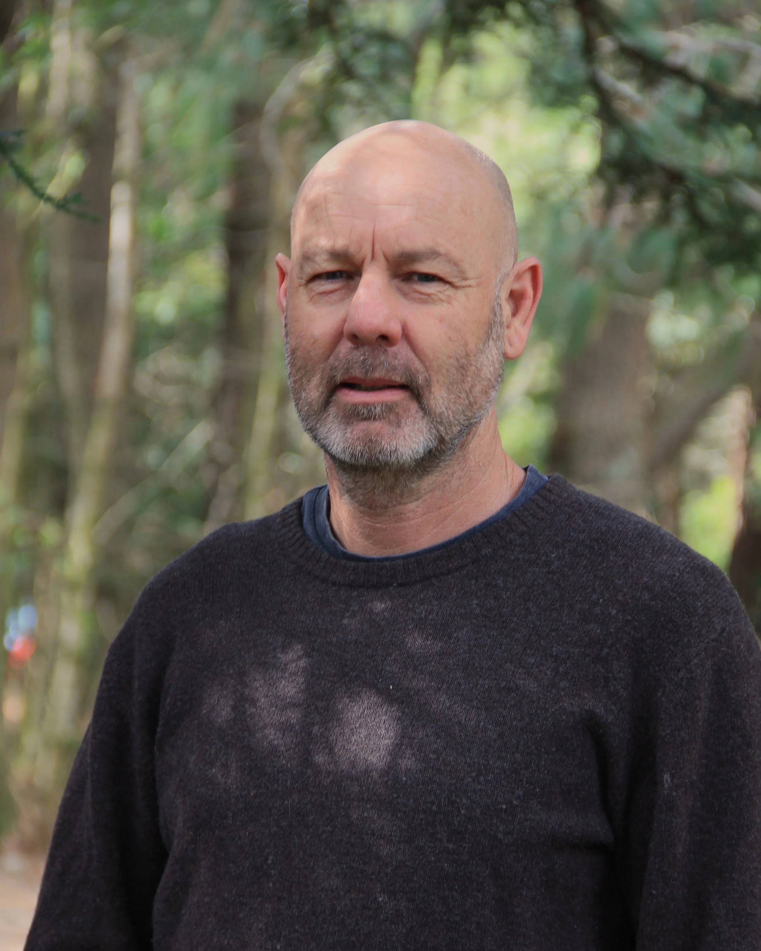 Close-up of a middle-aged man with a bald head, gray beard, and mustache, standing outdoors with trees and greenery in the background.