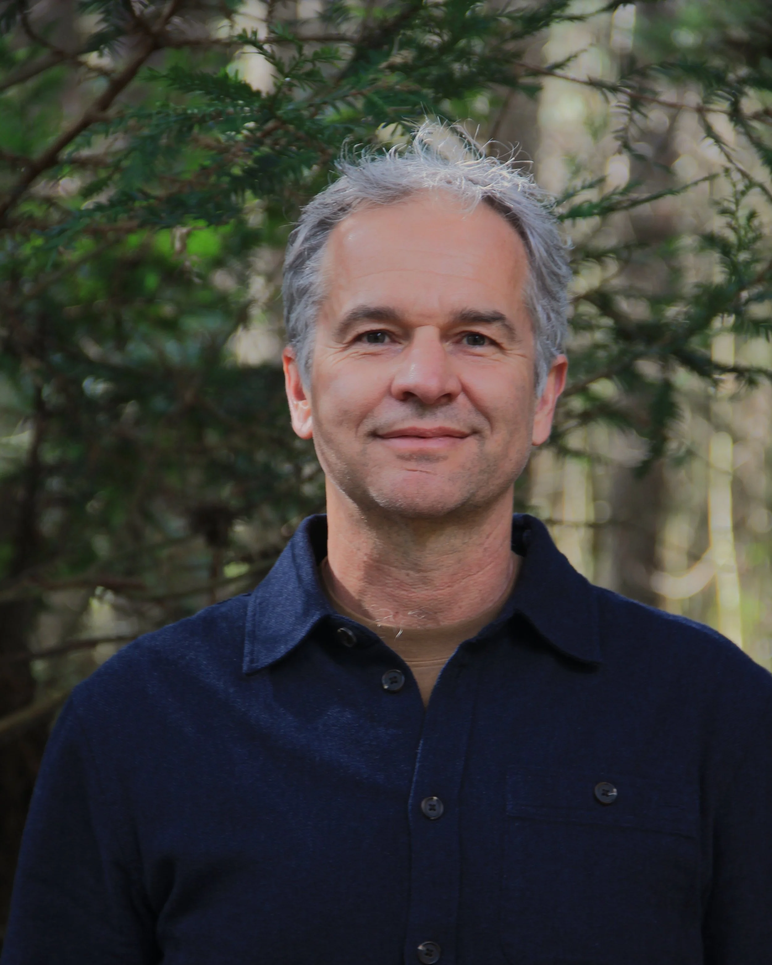 A middle-aged man with gray hair and light skin, wearing a dark blue collared shirt, standing in a wooded area with green foliage in the background.