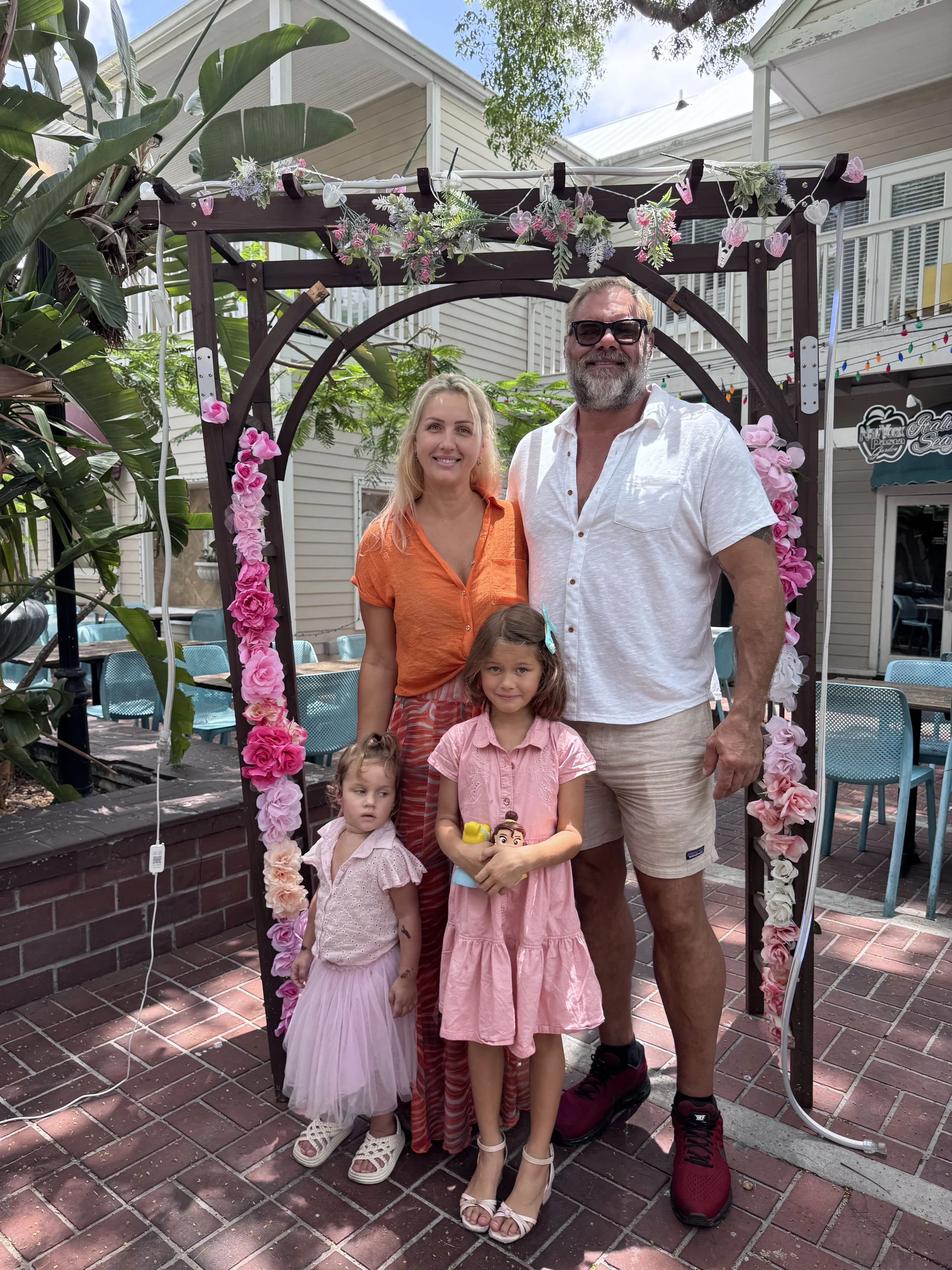 A family of five standing under a flower-decorated archway outdoors. Two adults, a man with a beard and sunglasses, and a woman, along with three young girls, pose for the photo. The scene is set on a brick patio with outdoor seating and lush green plants in the background.