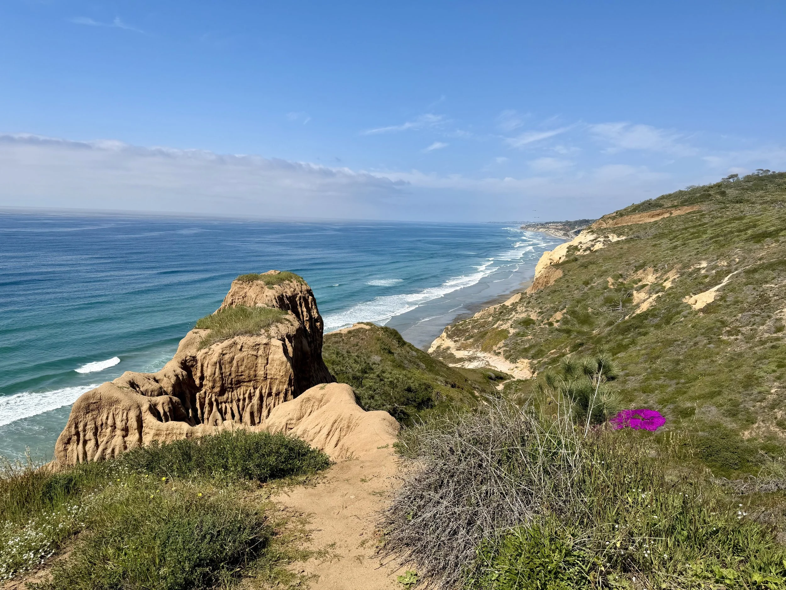 Scenic coastal view with sandy cliffs, green vegetation, and ocean waves under a blue sky with clouds.
