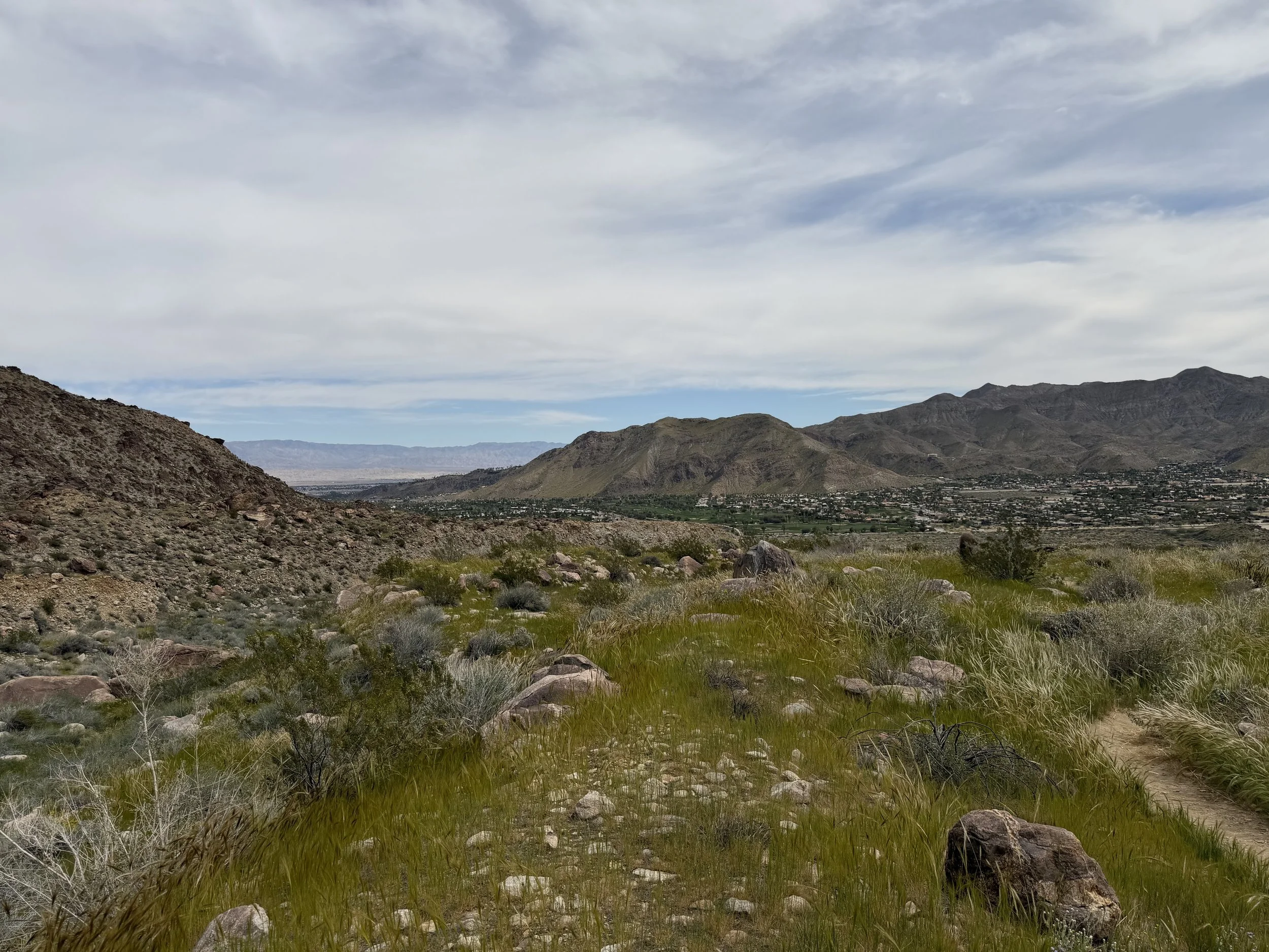 A landscape view of desert mountains under a partly cloudy sky, with rocky terrain and sparse vegetation in the foreground.