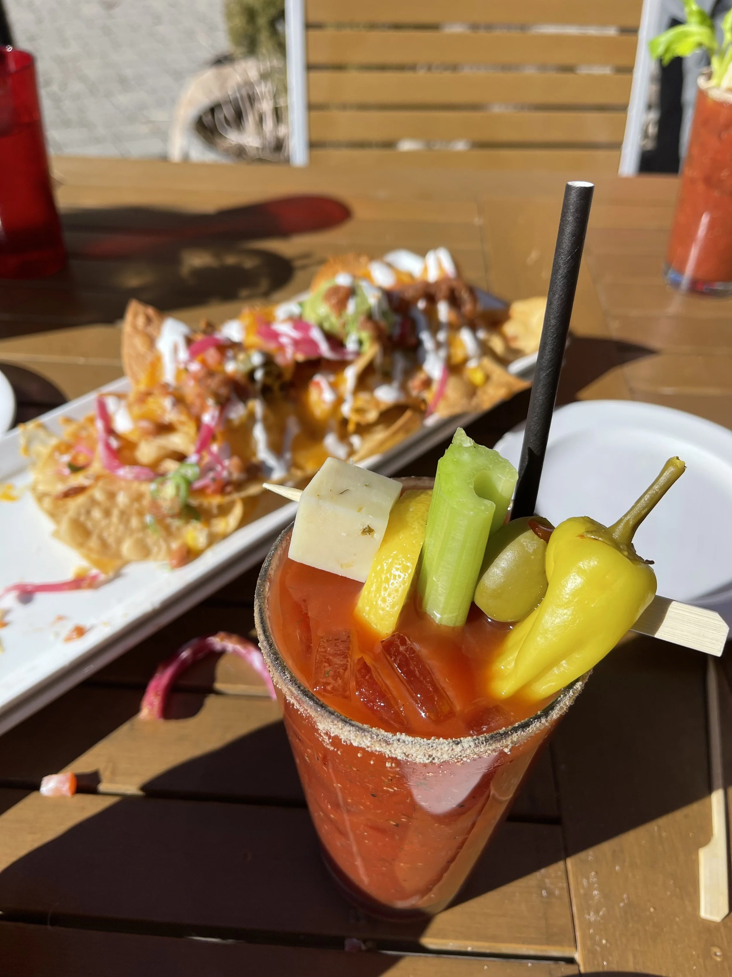 A spicy Mexican cocktail garnished with an assortment of vegetables and cheese, served in a salt-rimmed glass on a wooden table.