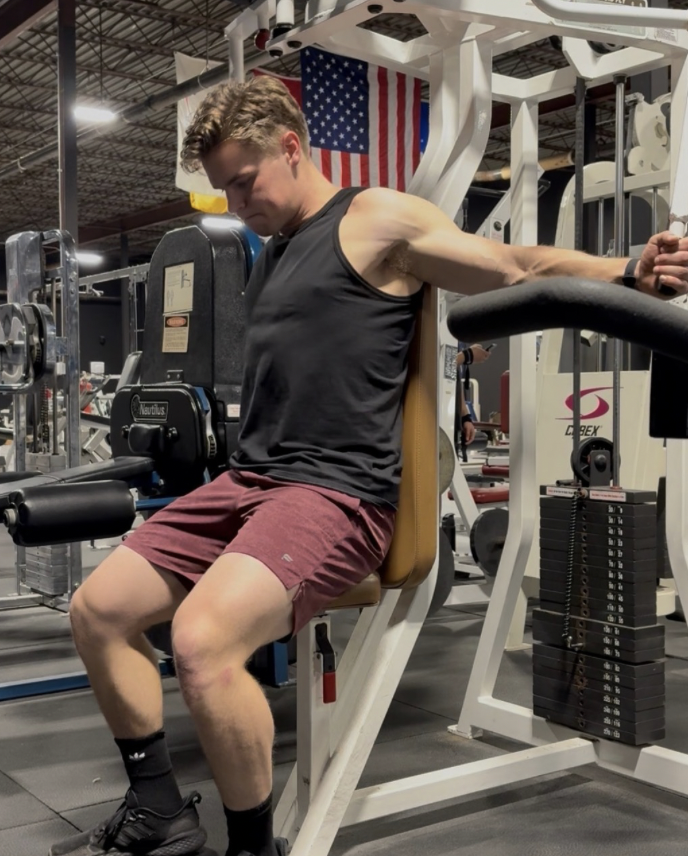 Man in a gym sitting on a weight training machine with American flag in background.