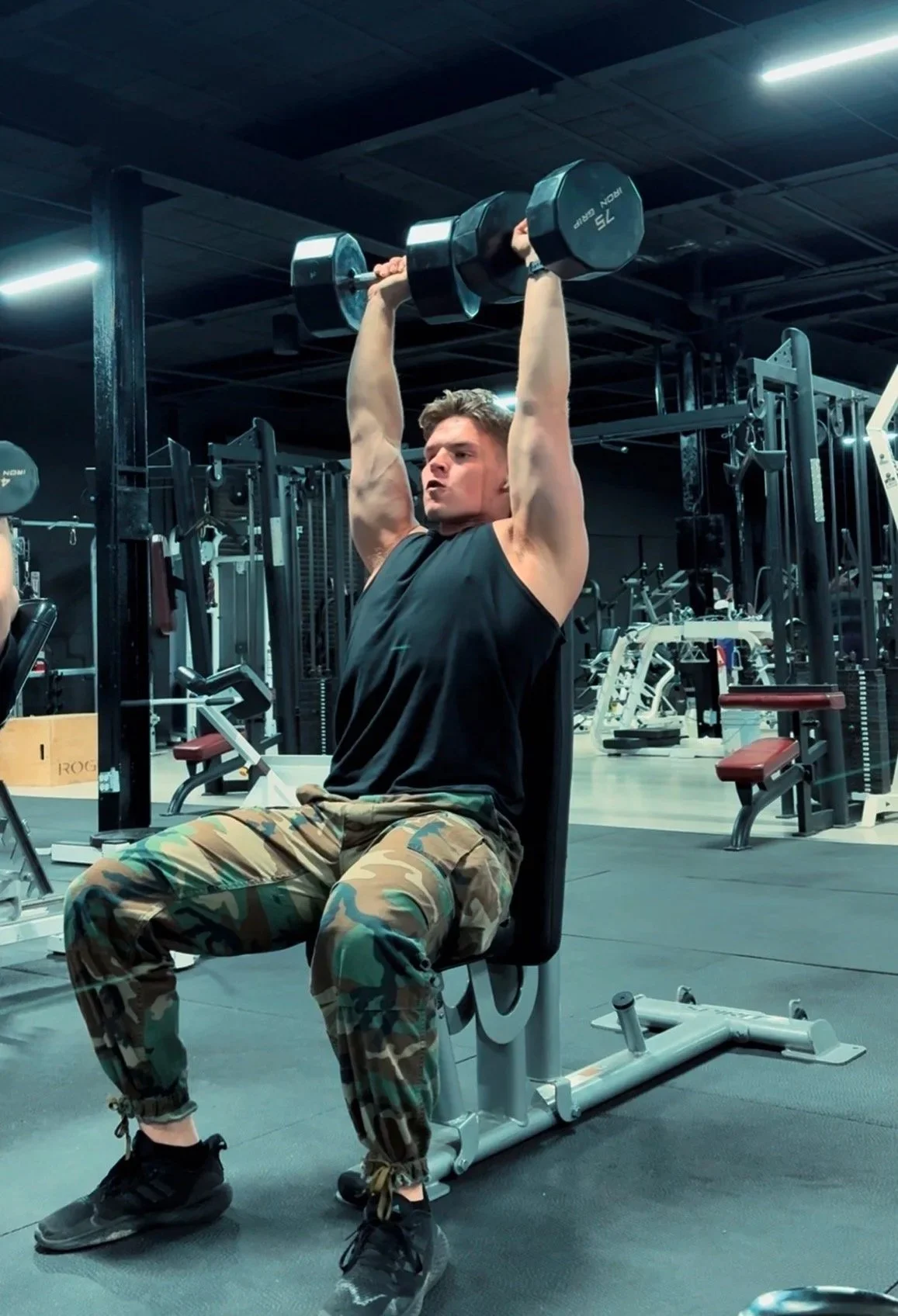 A man in camouflage pants and a black sleeveless shirt is seated on a workout bench in a gym, lifting a barbell with weights overhead, with gym equipment visible in the background.
