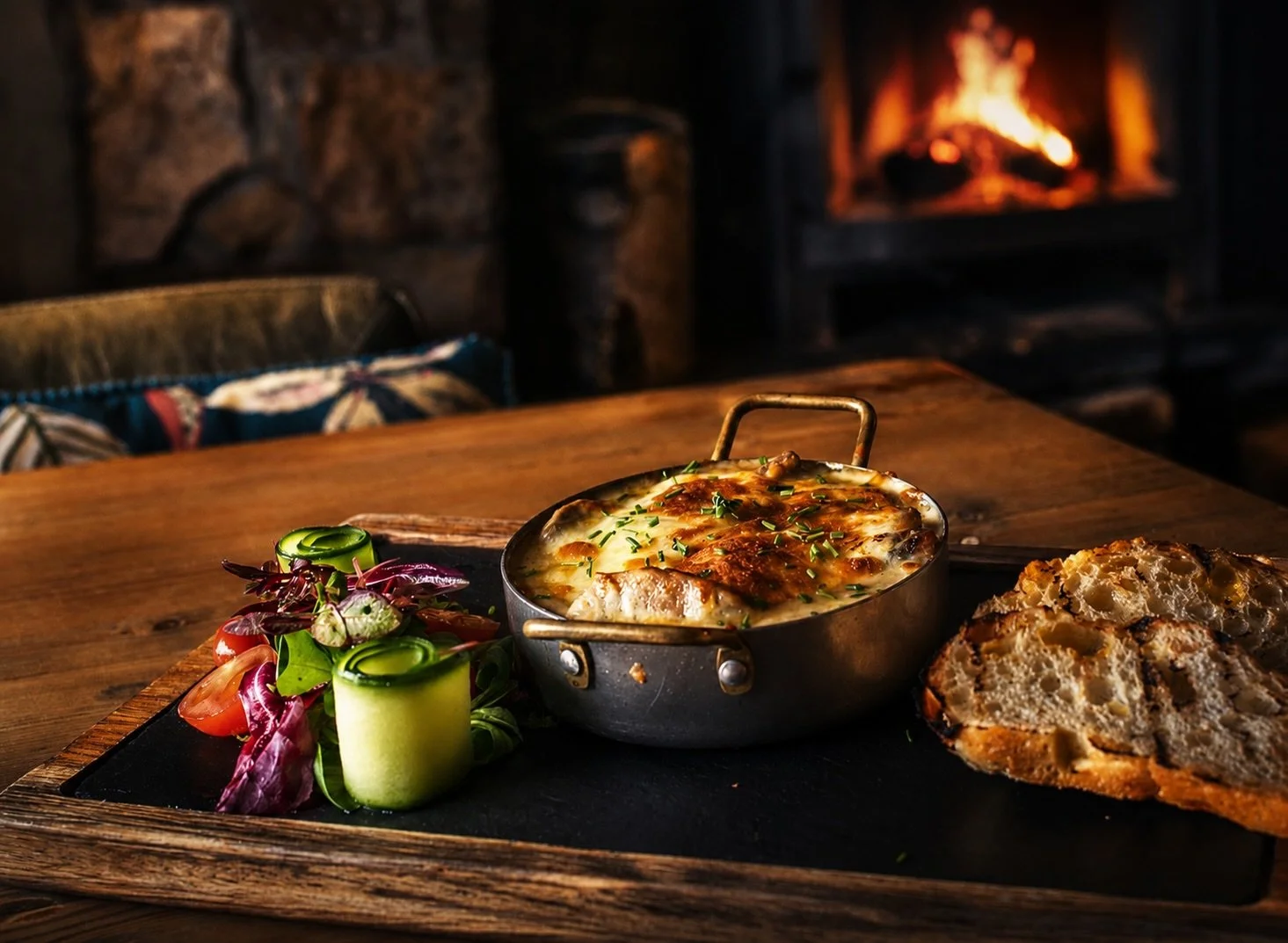 A rustic wooden table holding a dish of baked cheesy casserole with chopped herbs, a slice of bread, and a colorful salad with cucumbers, tomatoes, and microgreens. A cozy fireplace glows in the background.