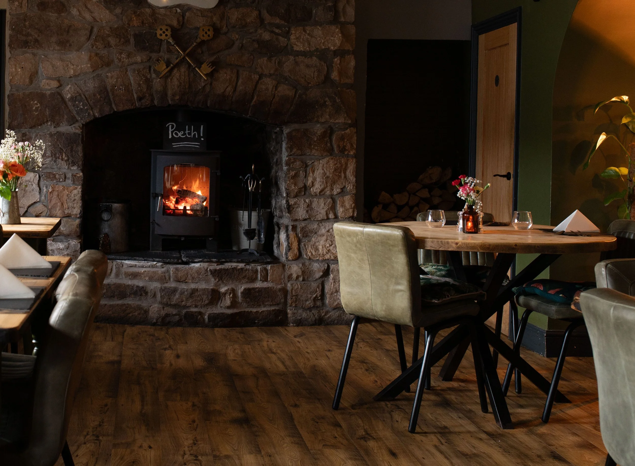 A cozy restaurant interior with a large stone fireplace and a wood-burning stove, decorated with a sign that says 'Poeth!', and tables set with flowers, glasses, and napkins. The atmosphere is warm and inviting.