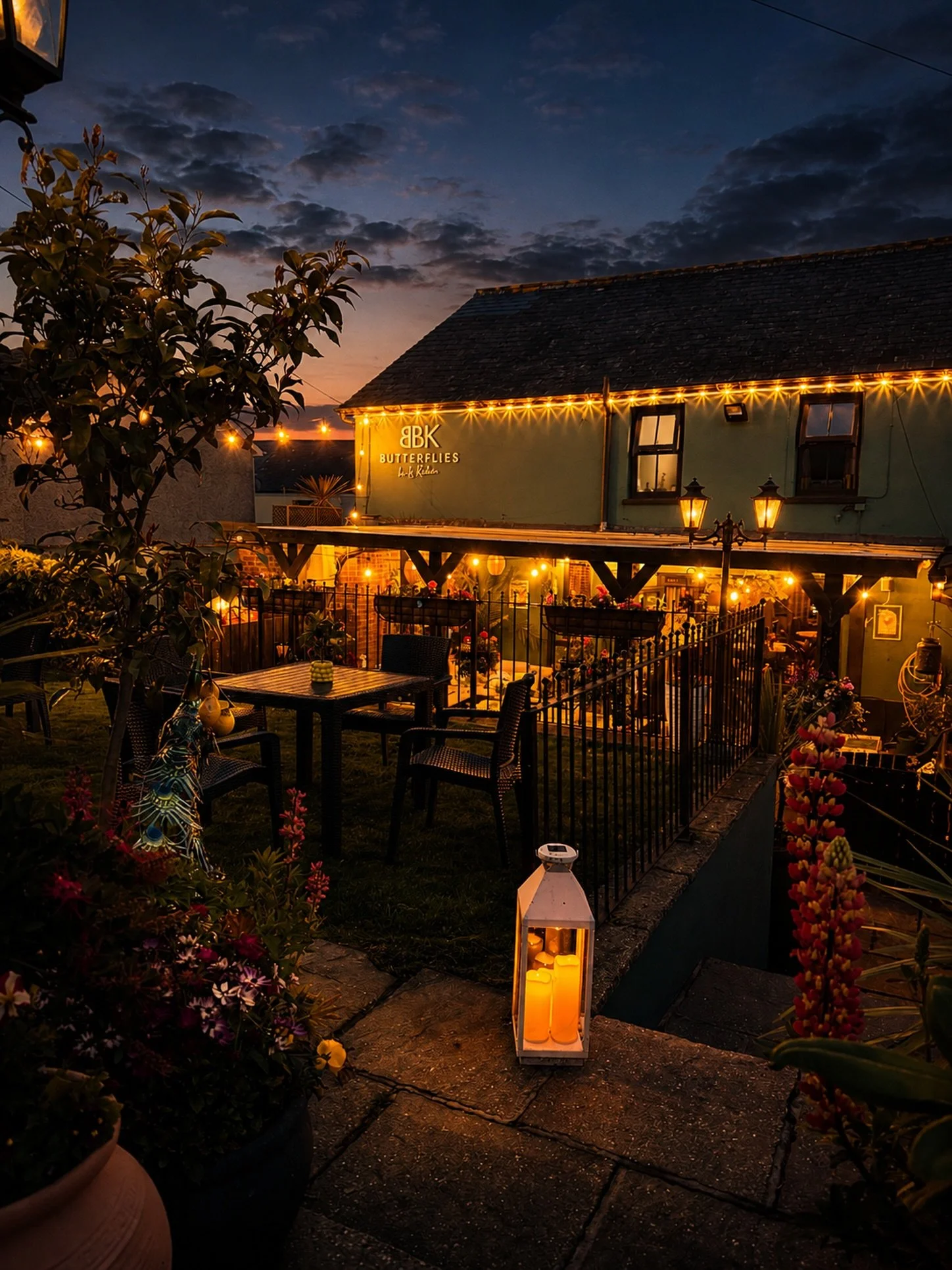 A cozy outdoor patio of a restaurant named 'Butterflies' at dusk, decorated with string lights and candles, featuring tables and chairs, surrounded by flowers and greenery.
