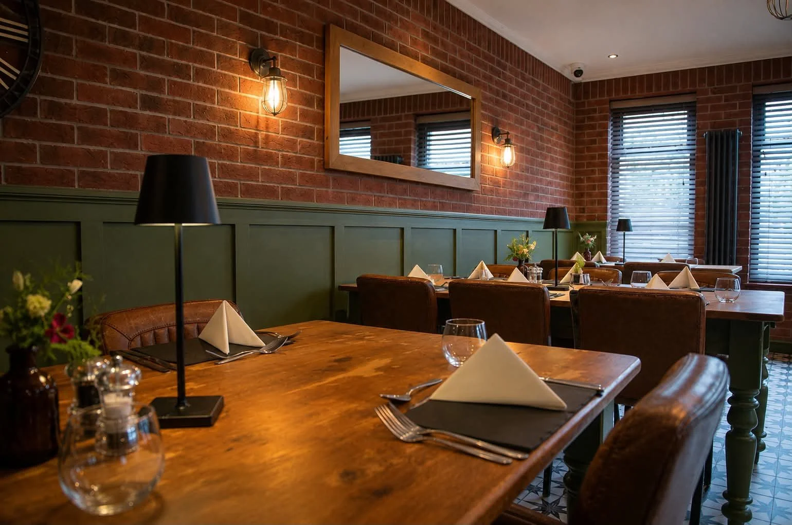 Interior of a cozy restaurant with brick walls, wooden tables, leather chairs, and large windows with blinds.