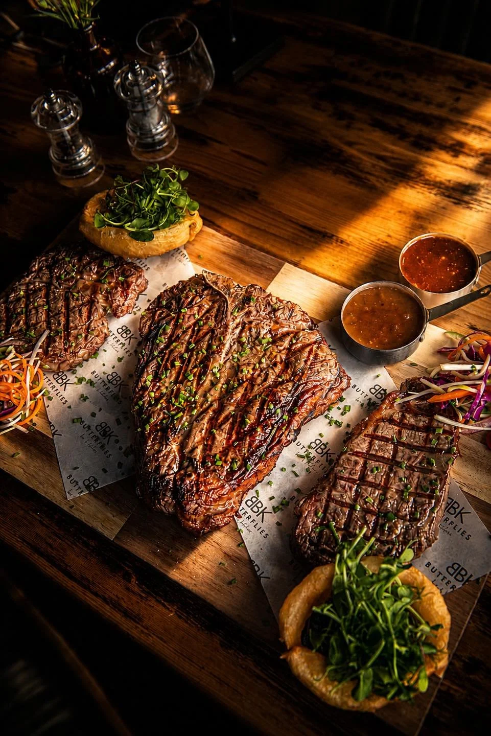 Barbecue ribs drizzled with barbecue sauce and garnished with chopped green onions, served with coleslaw, onion rings filled with microgreens, and two bowls of barbecue sauce on a wooden table.