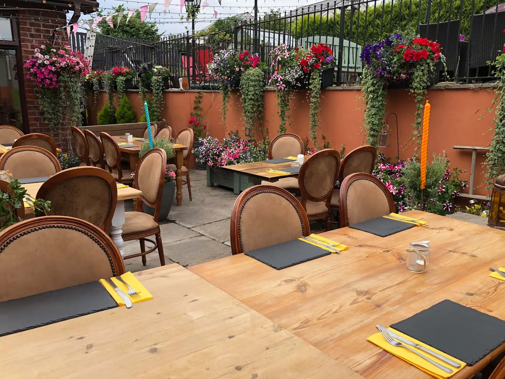 Outdoor dining area with wooden tables set with yellow napkins and black placemats, surrounded by chairs, decorated with colorful flowers, hanging pink and white bunting, and a fenced patio with plants and garden decor.