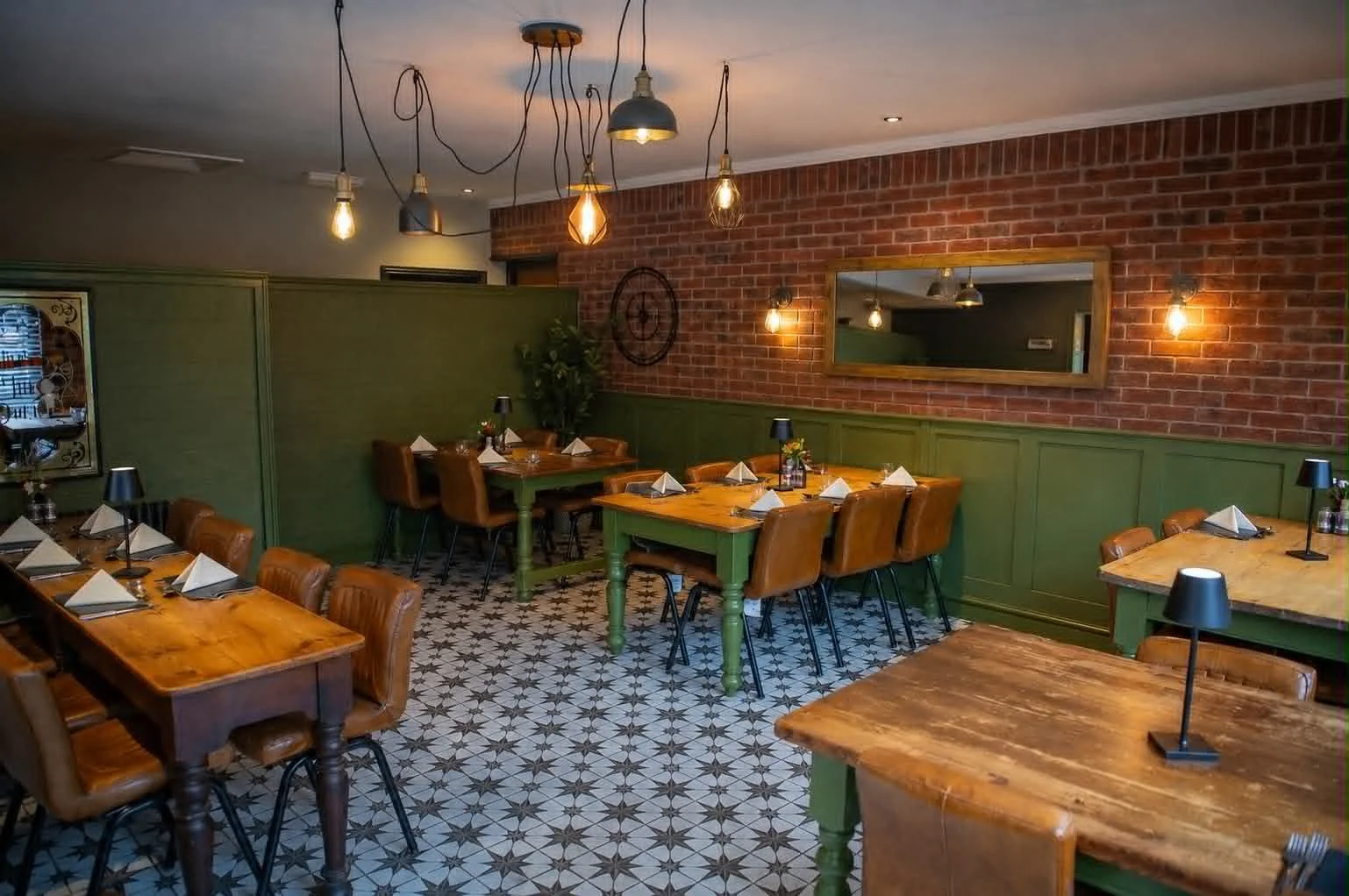 Interior of a cozy restaurant with wooden tables and leather chairs. The decor features a brick wall, green wainscoting, hanging pendant lights, and a decorative mirror. The floor has black and white geometric tile pattern.