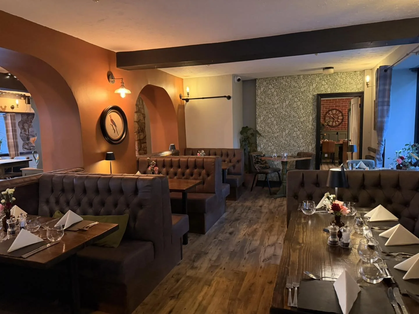 Cozy restaurant interior with wooden tables set with white napkins, wine glasses, and small floral centerpieces. There are brown tufted booths and chairs, ambient lighting, and a patterned wall at the back. A doorway shows an adjacent room with a wall clock and brick accents.