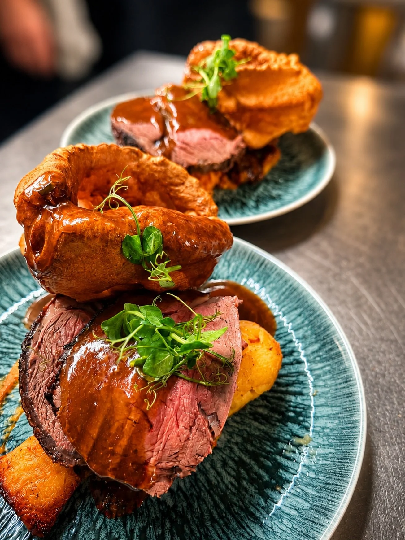 Close-up of a plated beef dish with a rich brown sauce, garnished with microgreens, served alongside roasted vegetables, on a textured blue-green ceramic plate.