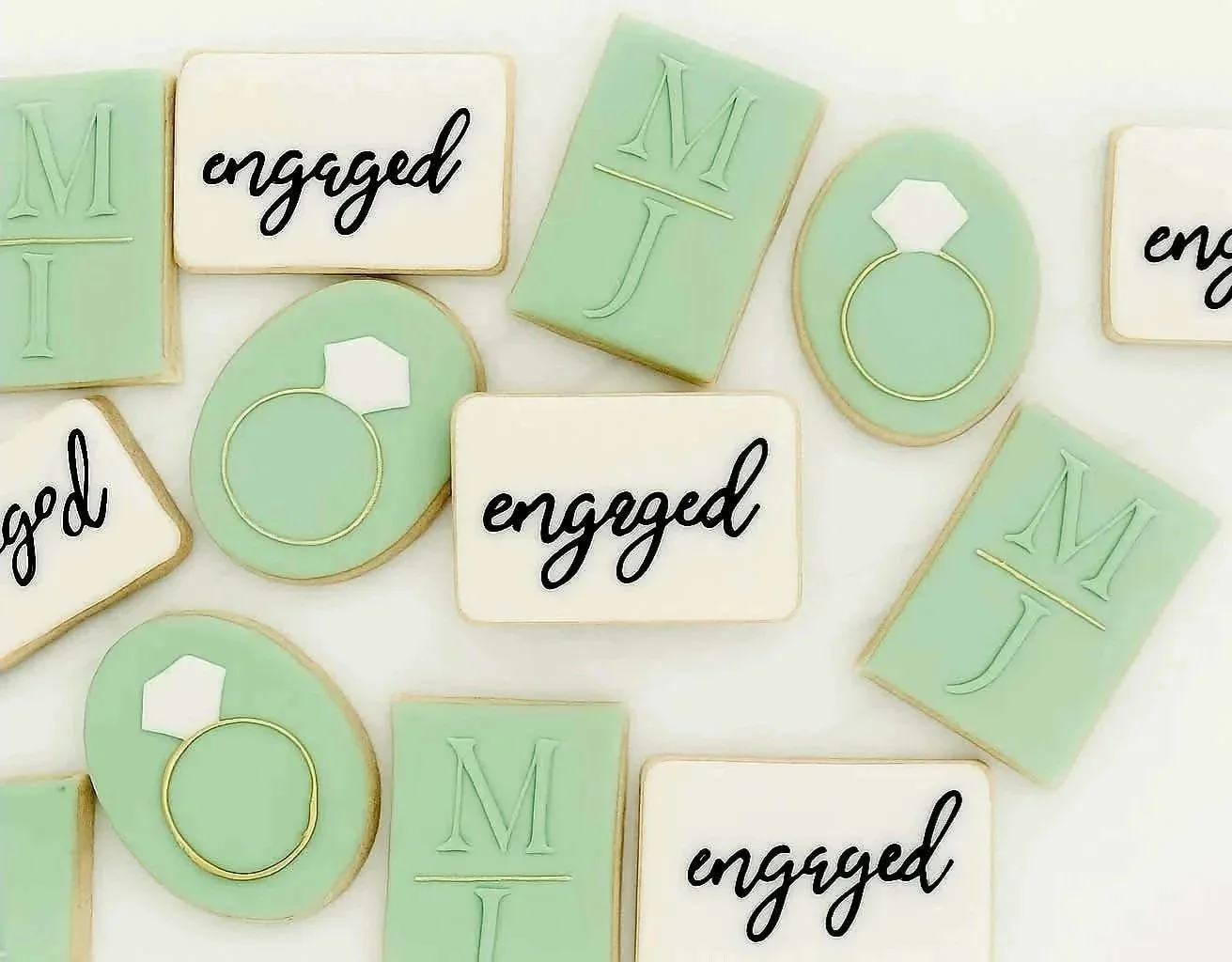 Assorted decorated cookies with engagement theme, featuring the words 'engaged' and initials 'M' and 'J,' along with ring designs in pastel green, white, and gold accents.