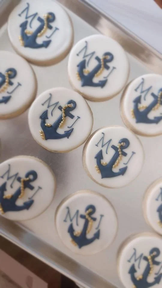 Cookies decorated with navy blue anchor and initials 'M' and 'J' along with gold accents on white icing, arranged on a tray.