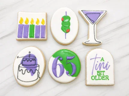 Six decorated cookies on a white surface. Purple and green birthday candles, green olives on a pick, a martini glass, birthday, the number 65 in purple with a greeen and white background and an arch cookie "A Tini Bit Older"