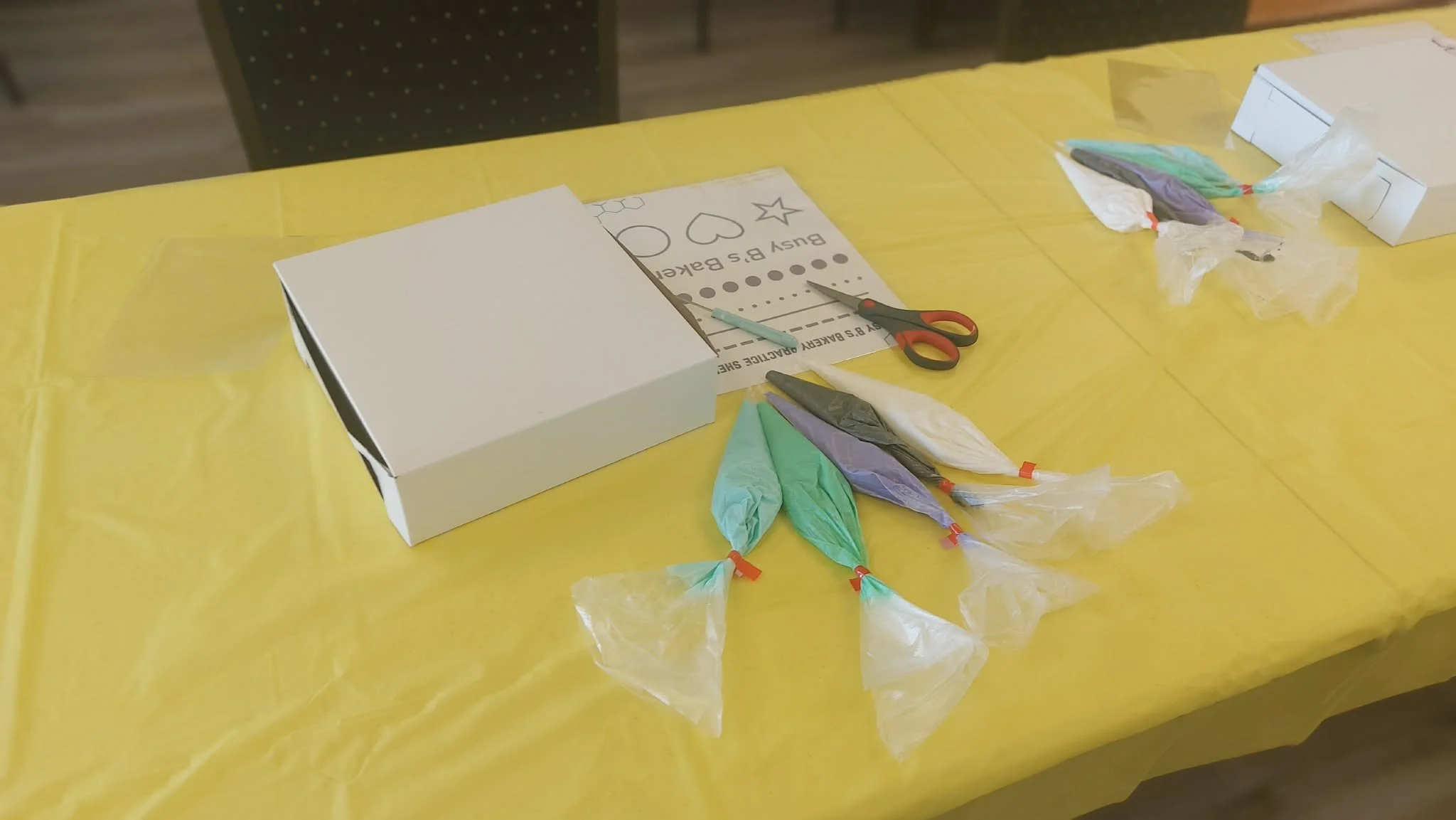 A table covered with a yellow tablecloth, with a white box, scissors, icing and a Busy B's Bakery practice sheet