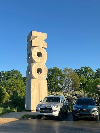Large concrete Zoo sculpture with geometric shapes, situated outdoors on a sunny day with parked cars in front and green trees in the background.
