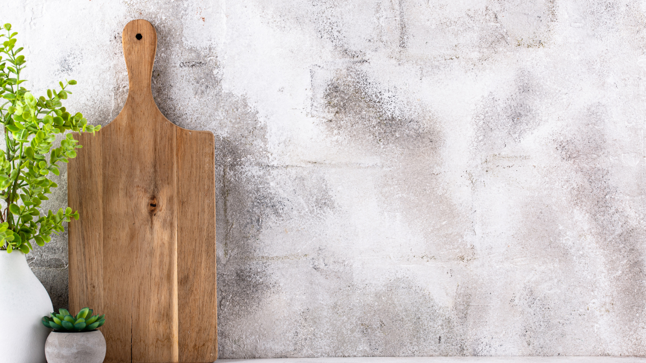 A wooden cutting board leaning against a textured light gray wall, with a white vase holding green leafy plants and a small succulent plant on the left.