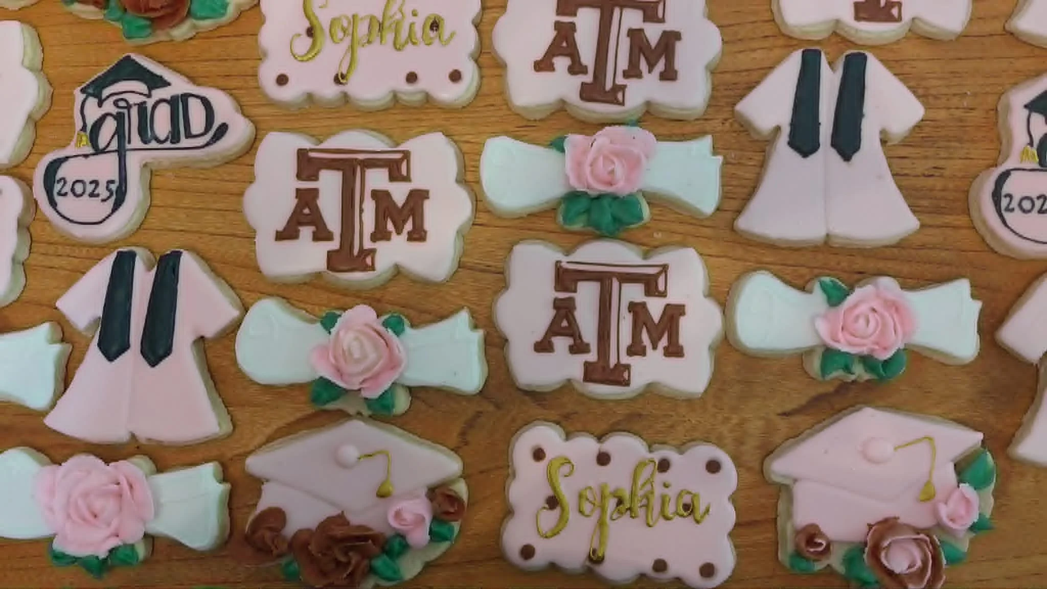 Collection of decorated cookies with themes related to Texas A&M University, featuring logos, flowers, and graduation symbols on a wooden surface.