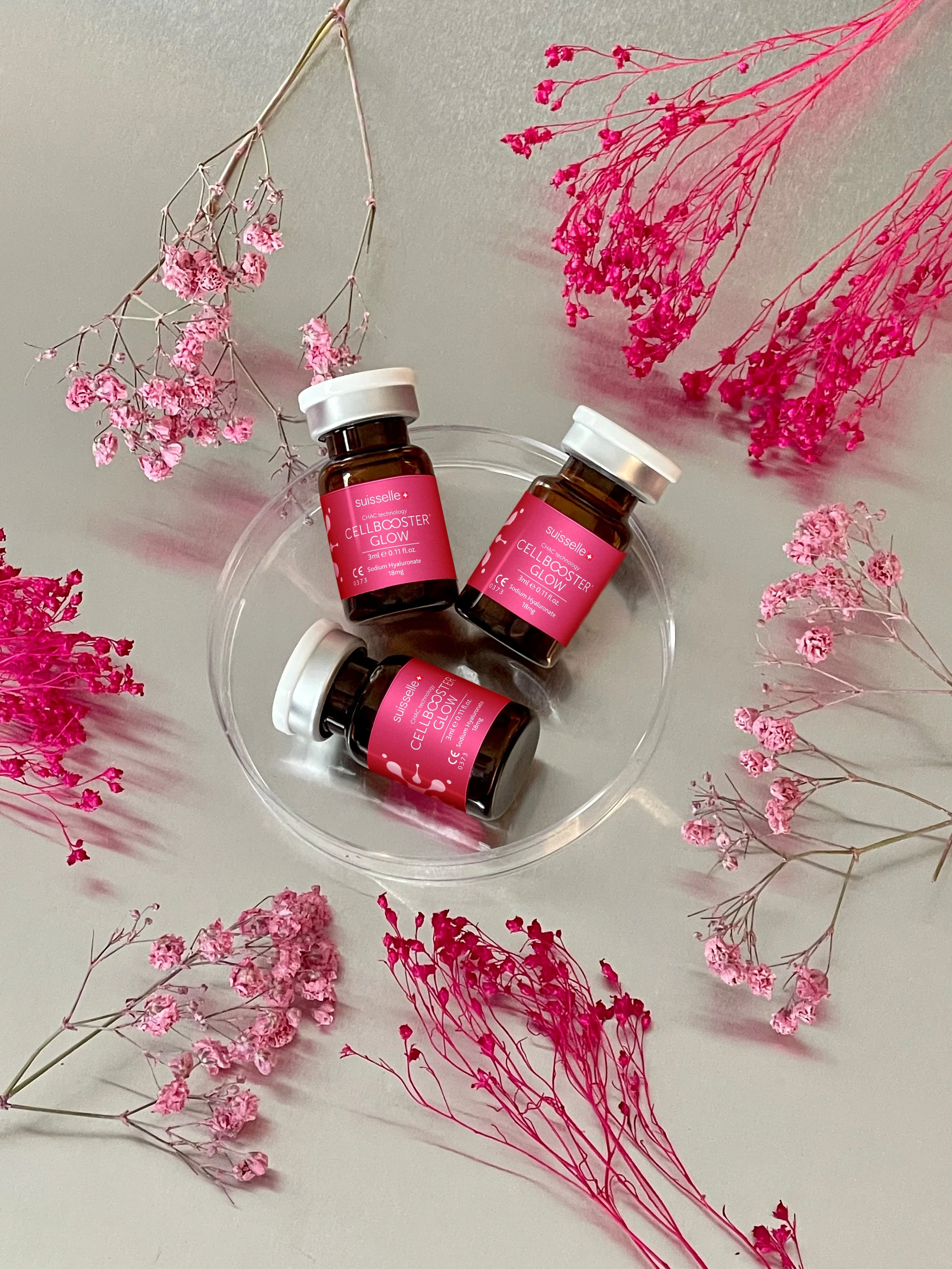Kolme pientä ruskean väristä lasipulloa, joissa on pinkit etikettit, suljettuna valkoisilla korkeilla. Pullojen etiketissä lukee 'CELL BOOSTER GLOW' ja ne on aseteltu muoviseen tarjottimeen. Pullojen ympärillä on erilaisia pinkkejä ja fuksian värisiä kuivattuja kukkia levitettyinä ympärille.