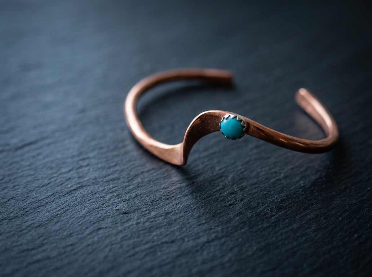 A copper cuff bracelet with a turquoise stone in the center, placed on a dark textured surface.