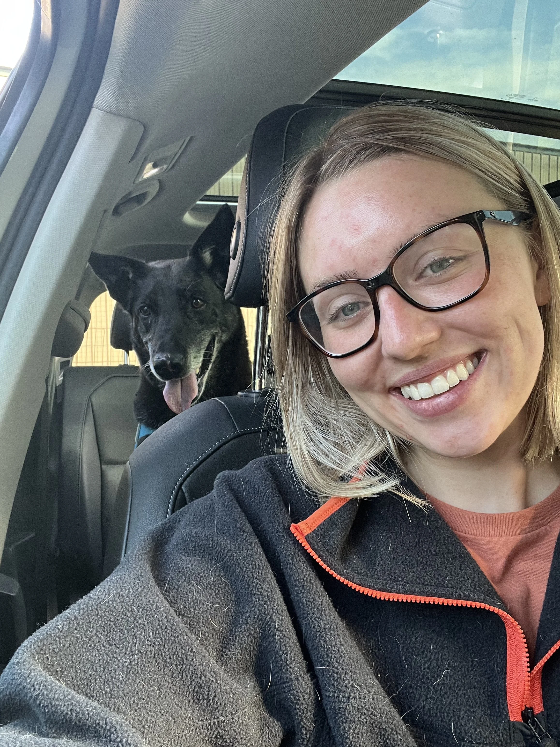 A woman with glasses and a smile, sitting in a vehicle with a black dog in the backseat, both looking towards the camera.