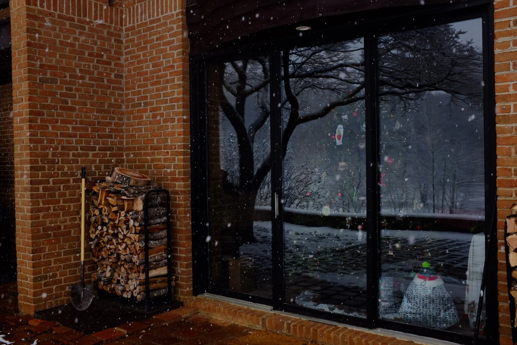 Snow falling outside a glass door with Christmas decorations visible through it. A stack of firewood is beside the door, and a snowman is inside, on the right side of the image.