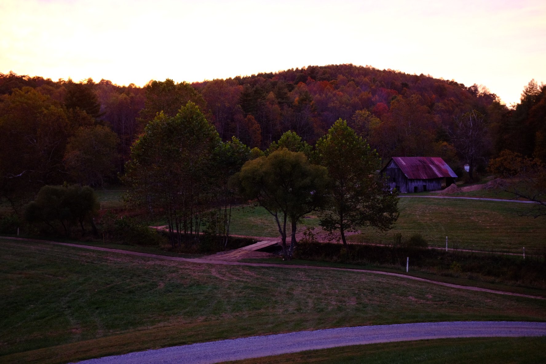 Sunset over a rural landscape with a hill covered in trees with fall foliage, a small wooden barn with a rusted metal roof, and winding dirt and gravel roads in the foreground.