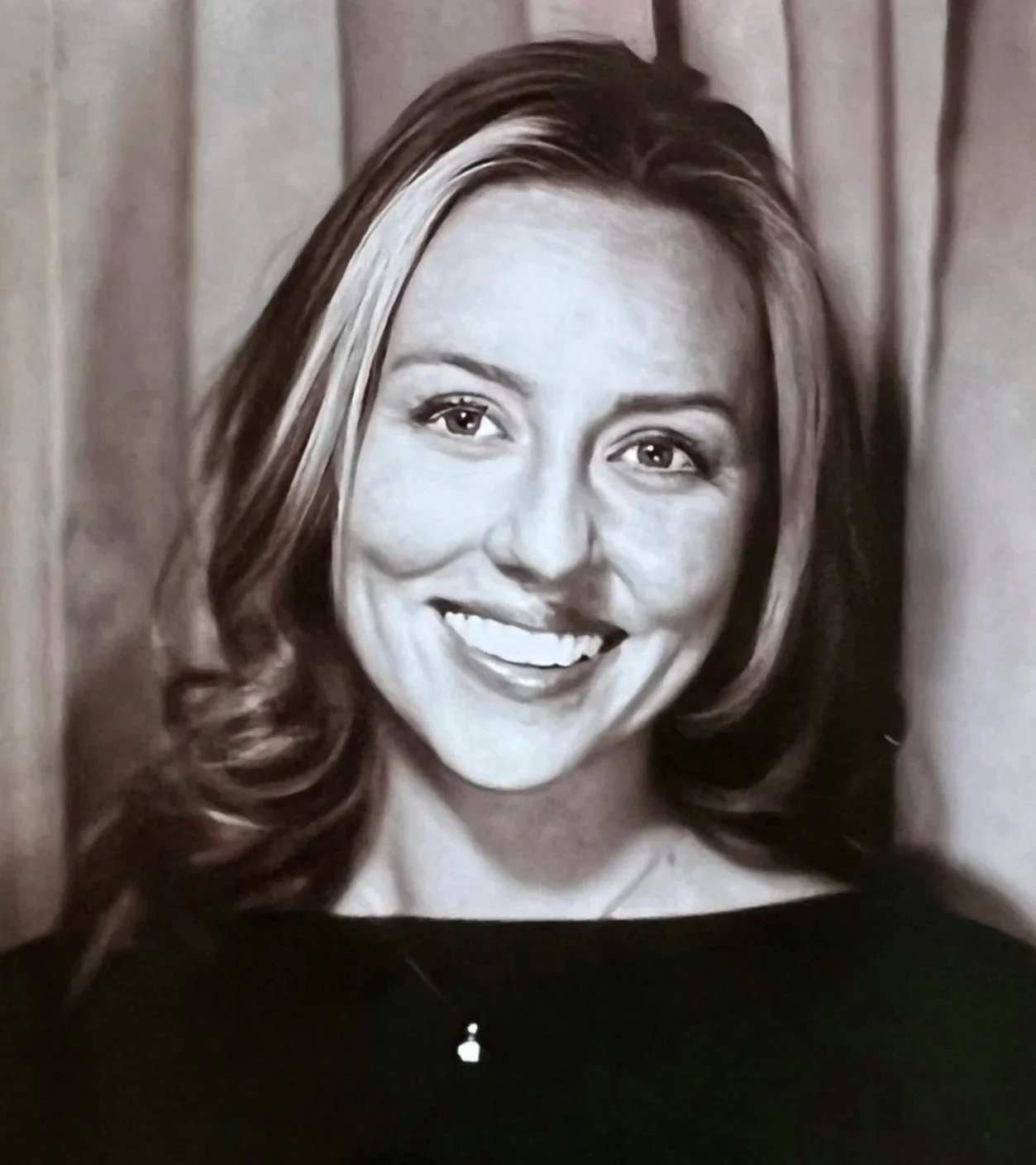 Black and white portrait of a smiling woman with long wavy hair, leaning against a wooden wall.
