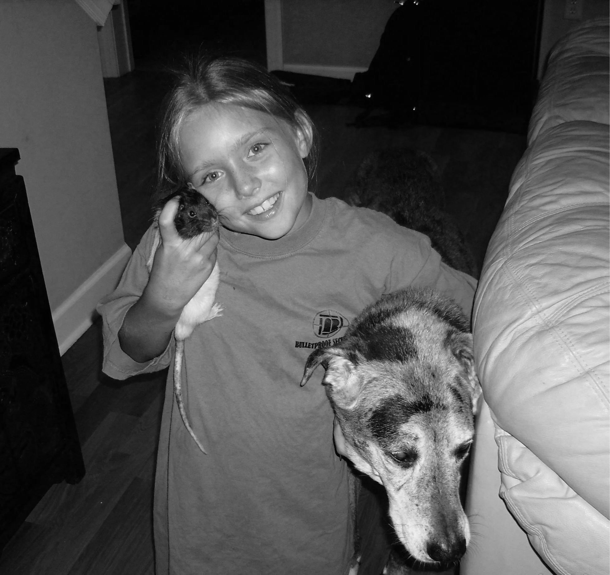 A young girl smiling while holding a guinea pig in her hand indoors, with a dog laying on the floor nearby and a couch to the right.