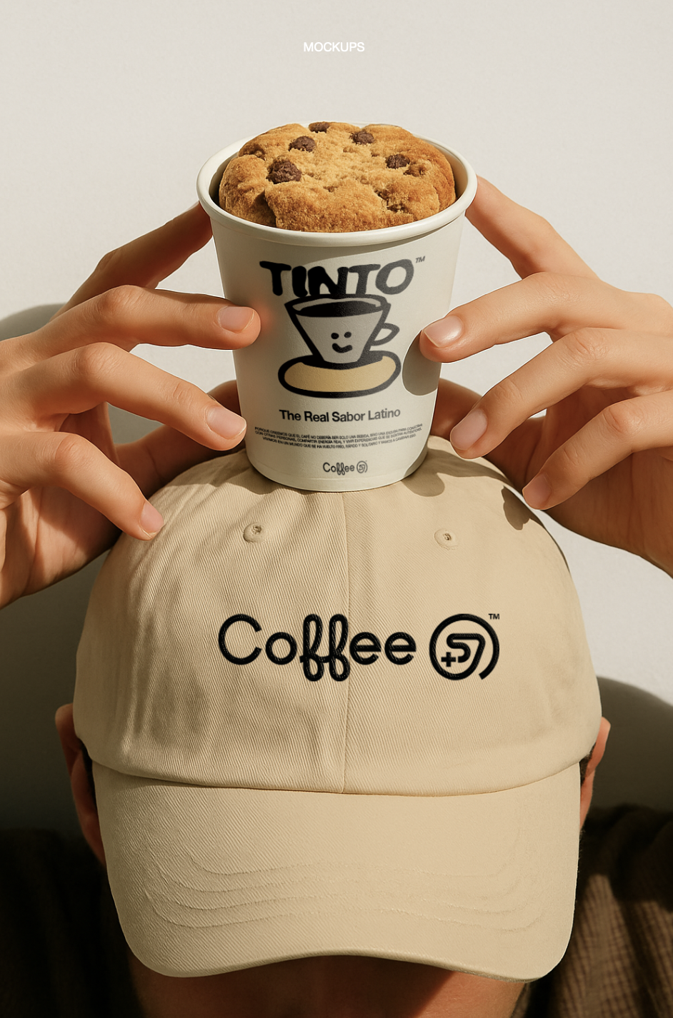 Person wearing a beige cap with 'Coffee+' logo holding a paper cup of chocolate chip cookie on top of their head with hands. The cup has a Tinto logo with a smiling coffee cup illustration and text reading 'The Real Sabor Latino.'