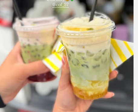Two hands holding cups of colorful bubble tea drinks with straws, one with a lemon slice and whipped topping, and the other with green and purple layers.