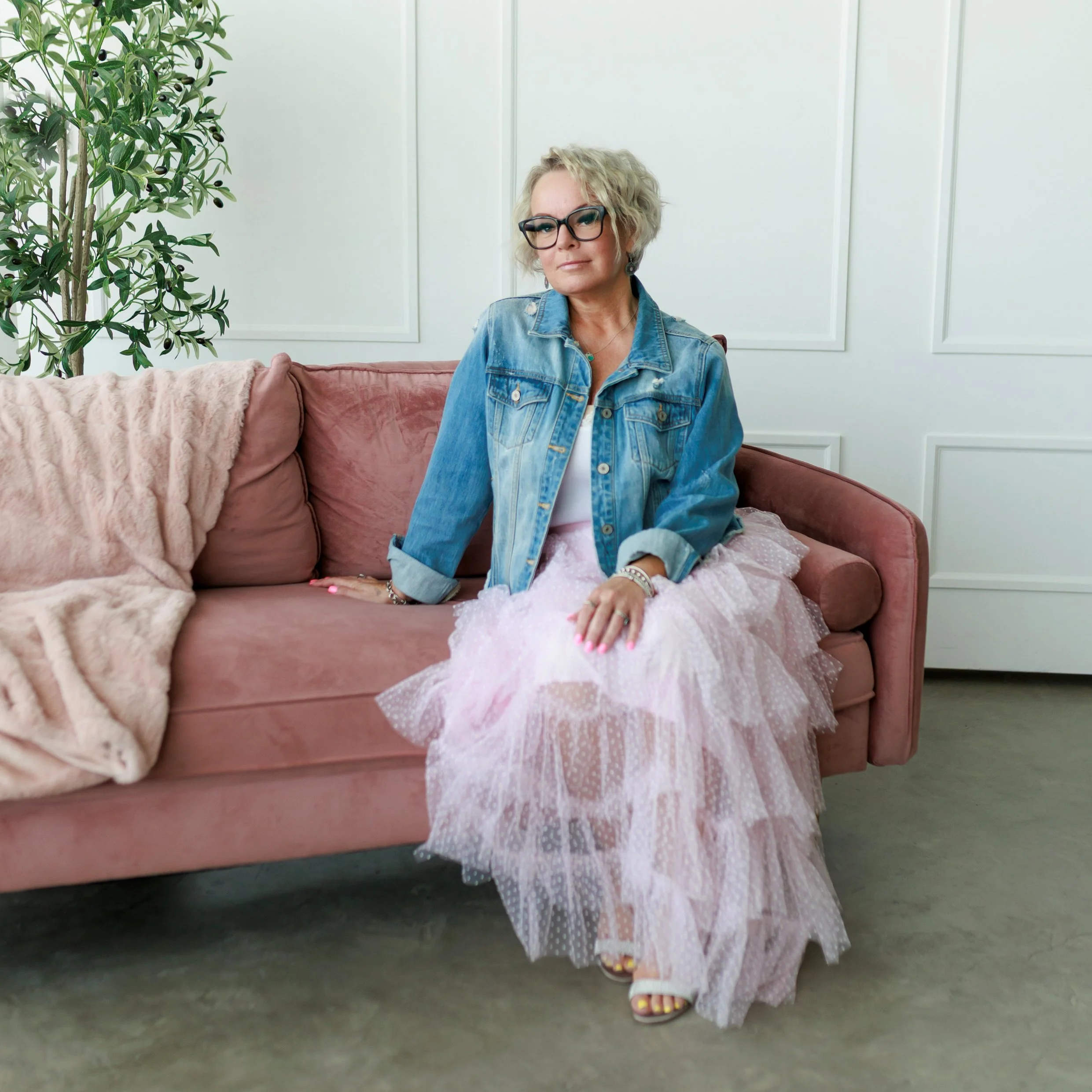 woman in pink tulle skirt and denim jacket with black glasses and blonde hair sitting on a pink couch