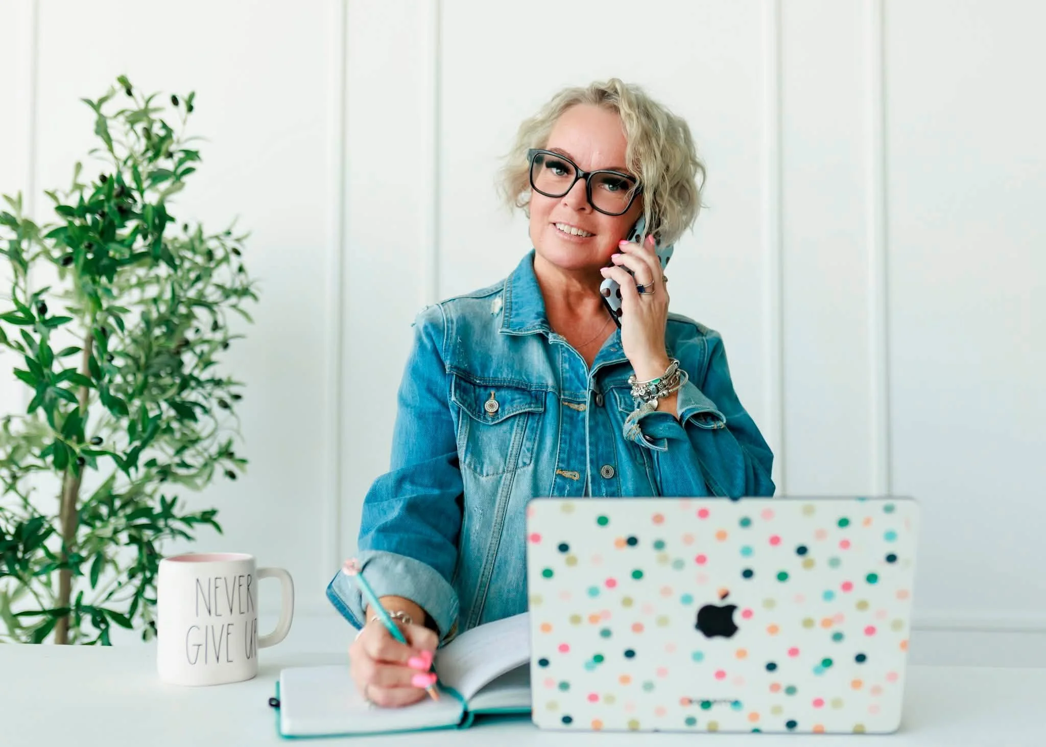 Woman with curly blonde hair, glasses, wearing a denim jacket, sitting at a white desk with a colorful laptop, talking on a cell phone, and writing in a notebook. On the desk, there is a white mug with text 'NEVER GIVE UP' and a small potted plant.