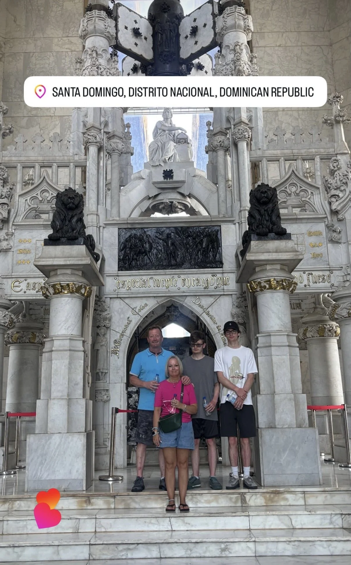 A group of five people standing on marble stairs in front of an ornate stone archway at a historical site in Santo Domingo, Dominican Republic. The archway features detailed carvings, columns, and statues, including two lion busts, with a statue of a seated woman above. There are small heart emojis at the bottom left corner of the image.