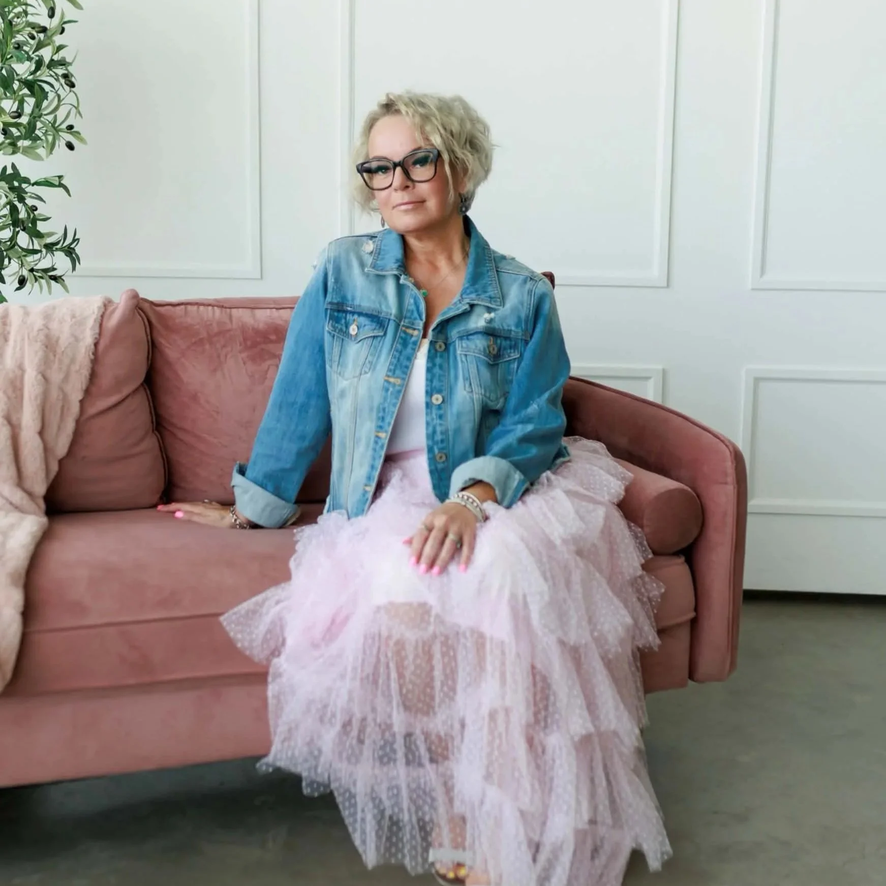 Woman with blonde curly hair, glasses, denim jacket, white shirt, pink tulle skirt, sitting on pink sofa, in a white room, next to green plant.