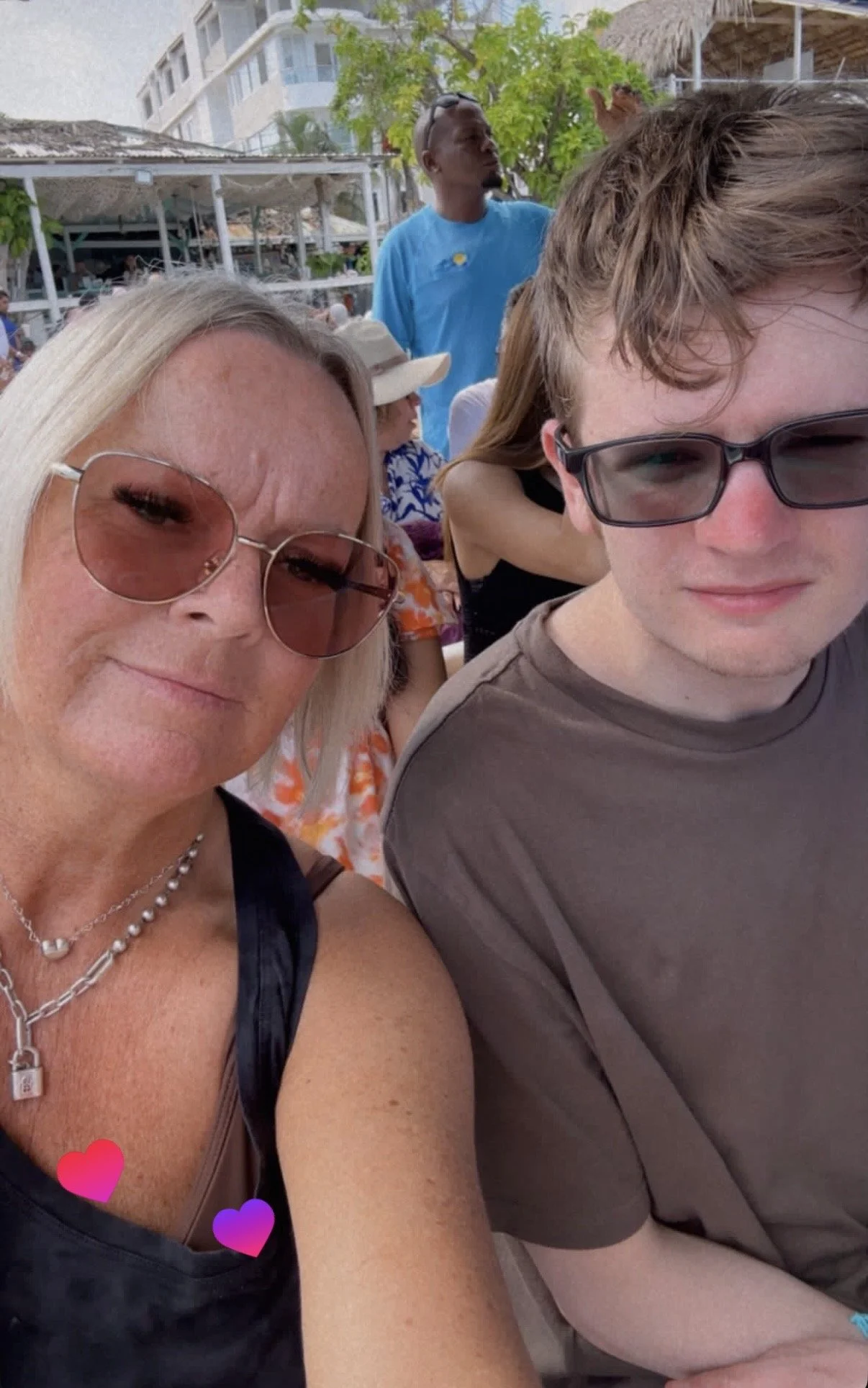 A selfie of two women and a young man outdoors at a crowded venue with buildings and trees in the background. One woman has blonde hair, sunglasses, and is wearing a black top with jewelry. The young man has light brown hair, glasses, and is wearing a gray T-shirt. Other people are visible behind them, including a man in a blue T-shirt and sunglasses, and a woman with a hat.