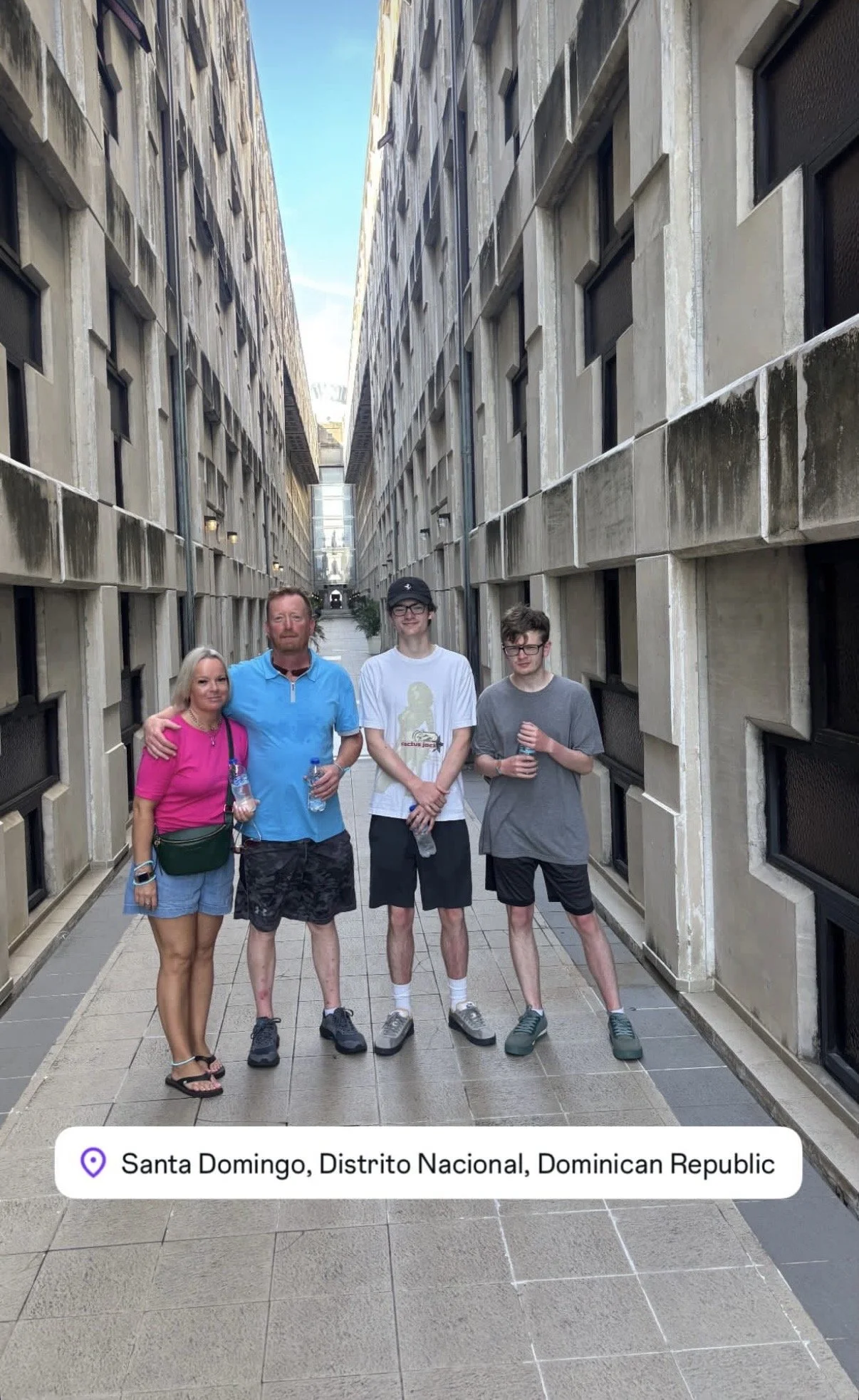 Four people standing together in a narrow alleyway between tall concrete buildings in Santo Domingo, Dominican Republic, with three holding water bottles, smiling at the camera.