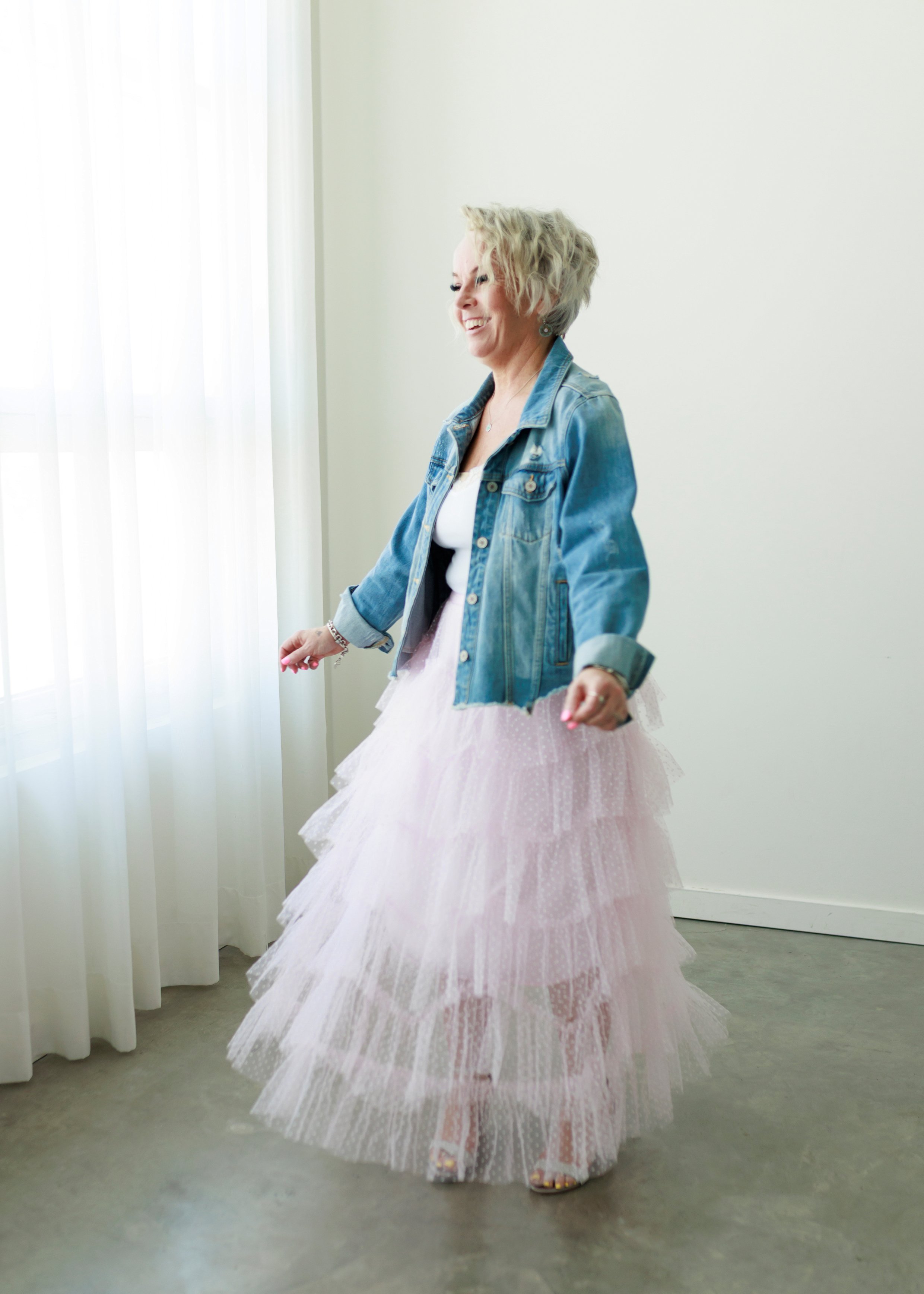 woman in pink tulle skirt and denim jacket lace trimmed tank and silver Steve Madden heels blonde waved hair smiling and laughing