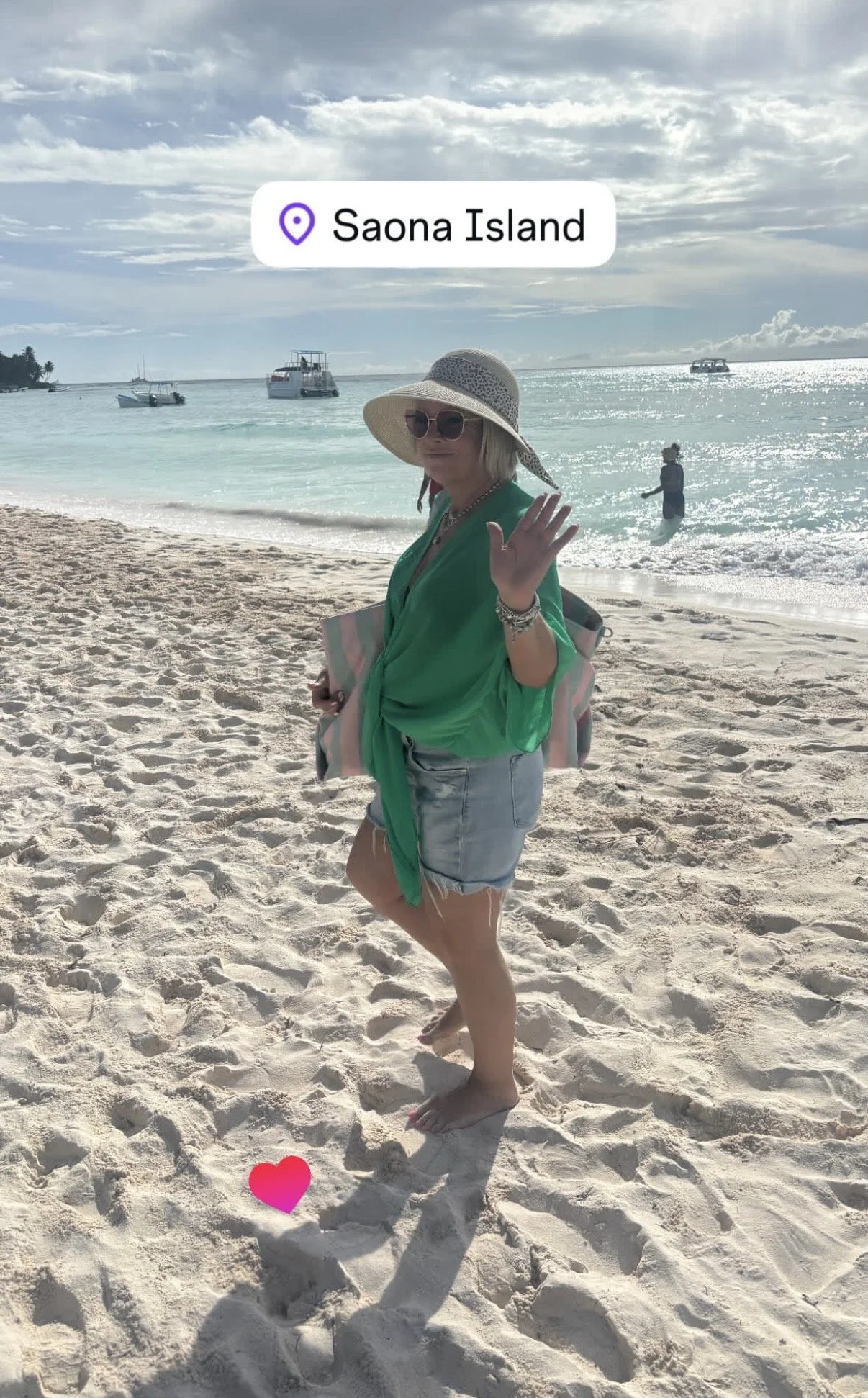 A woman on Saona Island beach, wearing a large sun hat, sunglasses, a green top, and denim shorts, waving at the camera. There are boats in the water and a person swimming in the background under a cloudy sky.