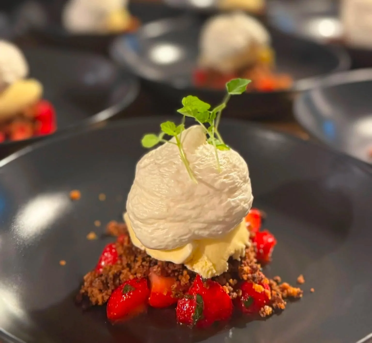 Un plat gourmet avec une quenelle de mousse blanche, garnie de petites feuilles vertes, sur un lit de fraises coupées en morceaux, dans une assiette noire.