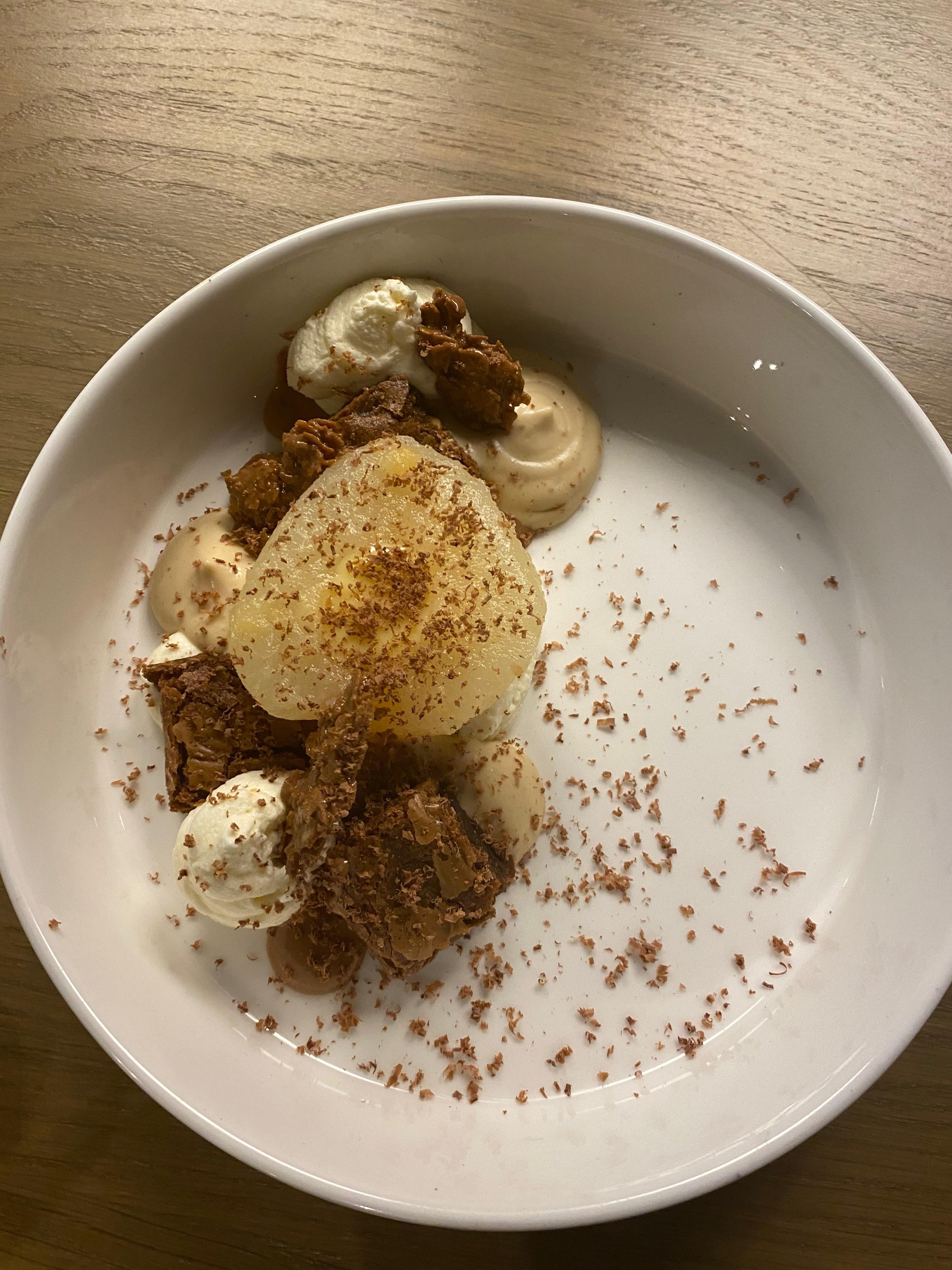 Dessert avec une portion de poire pochée, des morceaux de gâteau au chocolat, une quenelle de glace à la vanille et des morceaux de biscuit, décoré de copeaux de chocolat.