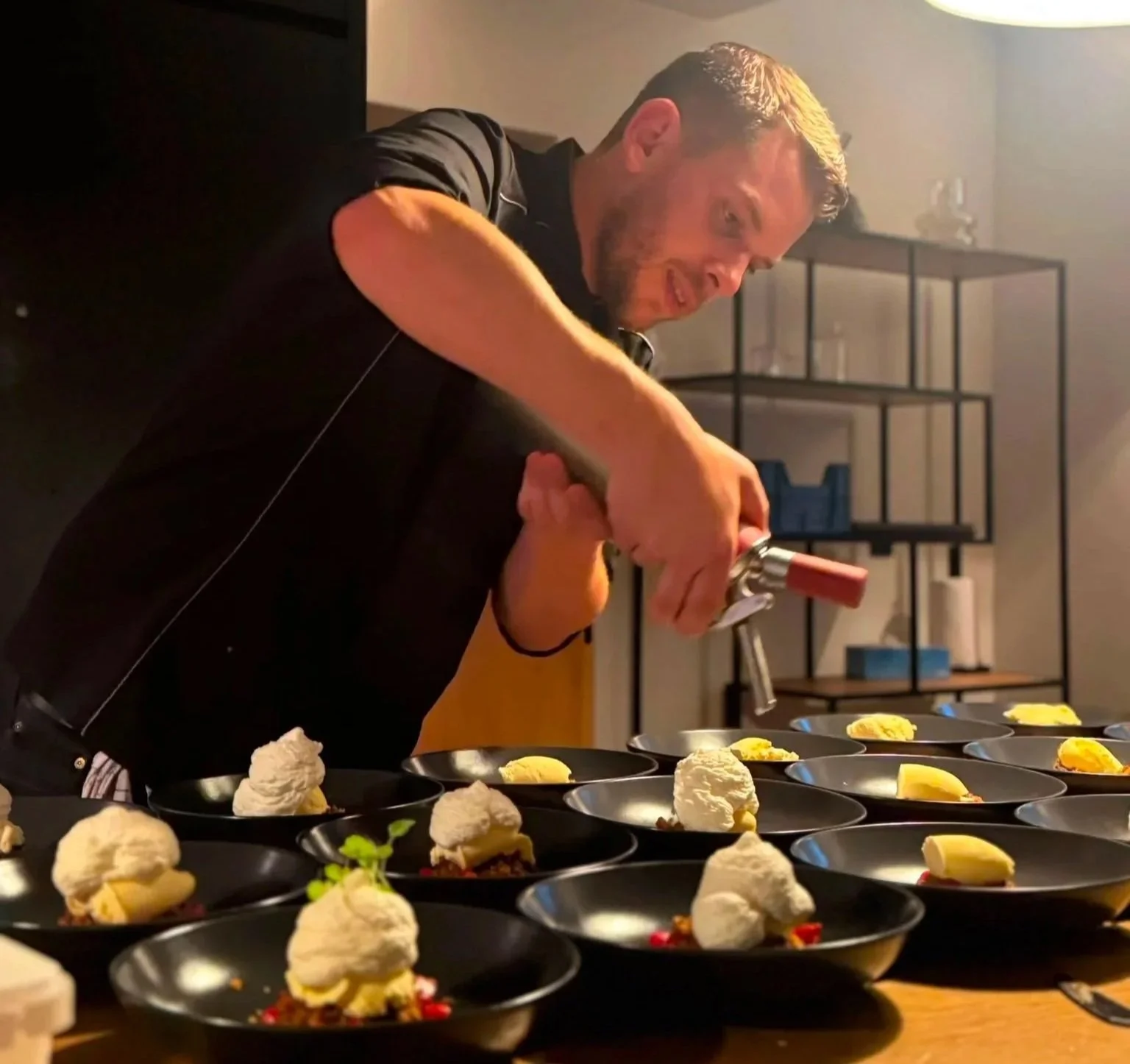 Un homme prépare des plats dans une cuisine, avec plusieurs assiettes devant lui contenant une portion de nourriture, possiblement un dessert ou une préparation culinaire.